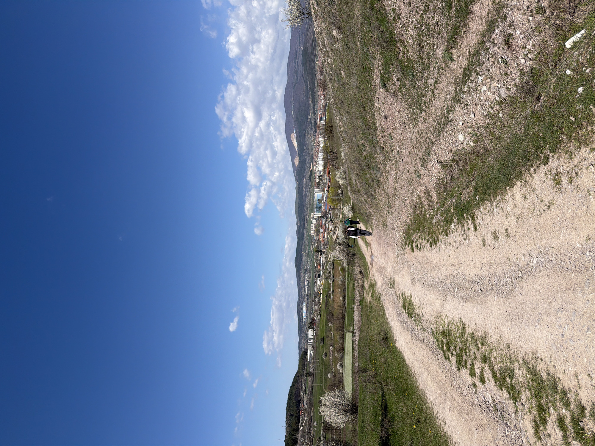 The image shows a rural scene near the city of Panagyurishte, Bulgaria. A dirt road winds through a landscape of grass and rocky terrain. A person or people, partially obscured, stand on the road. Beyond the immediate foreground, the city of Panagyurishte is visible, nestled against a backdrop of mountains under a sky with scattered clouds. The angle of the image suggests it was taken from a high vantage point, possibly a hillside overlooking the city.