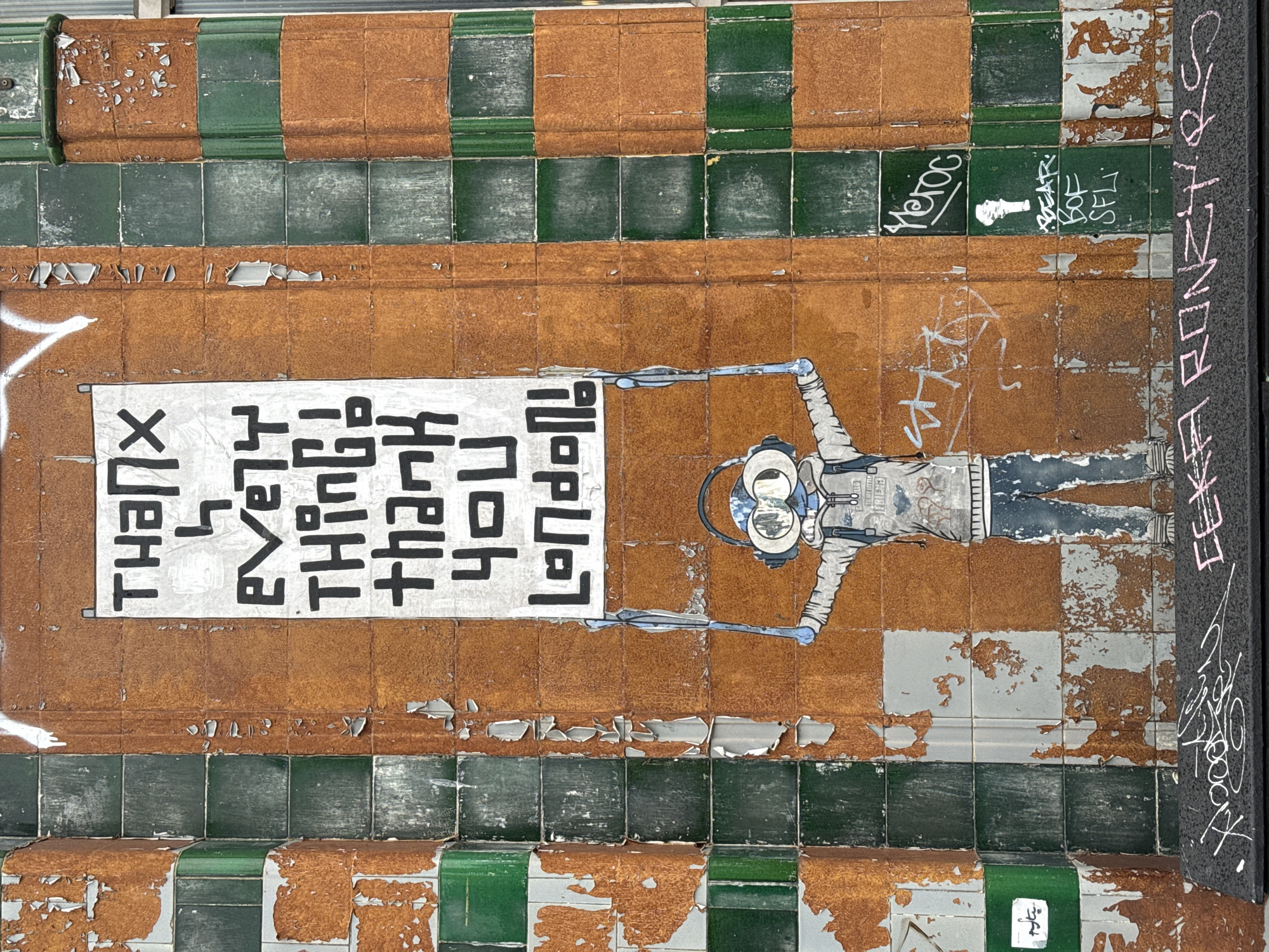 The image shows a section of tiled wall, with peeling paint and graffiti in a tunnel in the city of London. The tiles are green and brown, and a mural of an astronaut, seemingly holding a sign that is written backwards reads: THANK YOU FOR BEING FRIENDS.