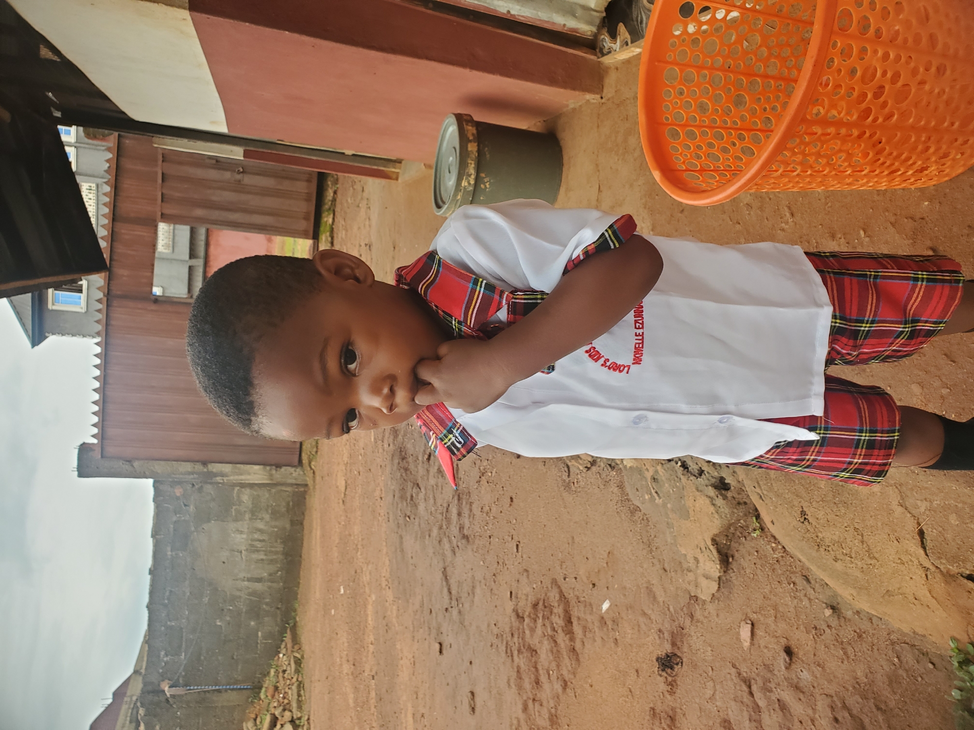 A young boy in a school uniform stands on reddish-brown dirt in Ukwu-Abwa, Nigeria. The uniform consists of a white short-sleeved shirt and red plaid shorts. The boy is looking off to the side, with his finger near his mouth. An orange plastic basket and a gray container are visible behind him near a reddish-brown building. A gray wall with a metal gate is to the left. The sky is overcast.