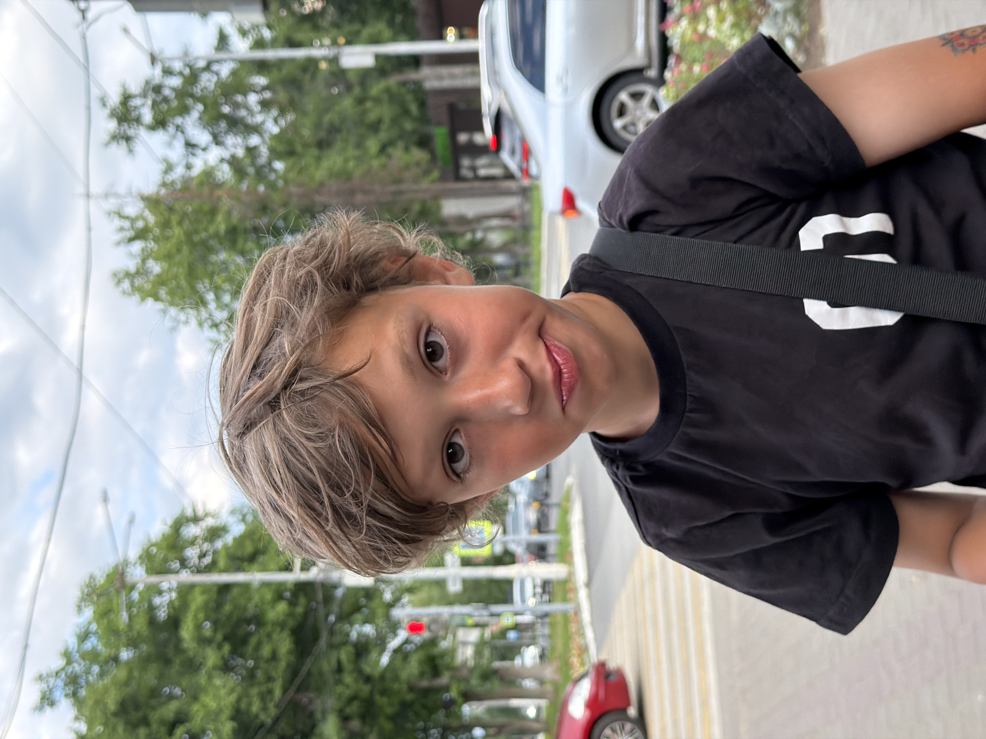 A young boy with tousled brown hair and expressive dark eyes looks directly at the camera, a slight pout on his lips. He is wearing a black t-shirt with a white graphic design on the front and a dark strap across his chest, indicating a bag or backpack. The background suggests an urban environment, with a blurred cityscape that includes trees, streetlights, and cars. A red car is visible on the left, and a silver car is parked on the right, with some flowering bushes in the foreground on the right. The scene appears to be outdoors, possibly on a sidewalk or street in Chaikovsky, Russia, indicated by the presence of a pedestrian crossing and street infrastructure. The lighting suggests it's daytime, with an overcast sky.