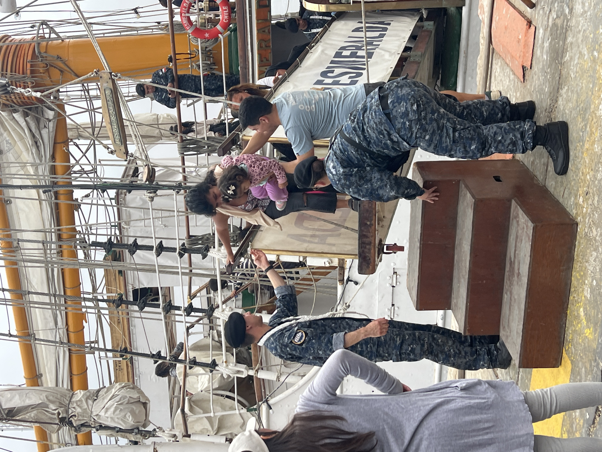 This photo was taken on the Esmeralda ship in Arica, Chile. The ship is a tall sailing vessel with a large mast and complex rigging. A Chilean naval officer in navy camouflage uniform guides a family: a man in a blue shirt, a woman, and a small child wearing a pink outfit up a small set of steps onto the ship. Other crew members and visitors are present. A life preserver with "ESMERALDA CHILE" printed on it, is present on the ship.