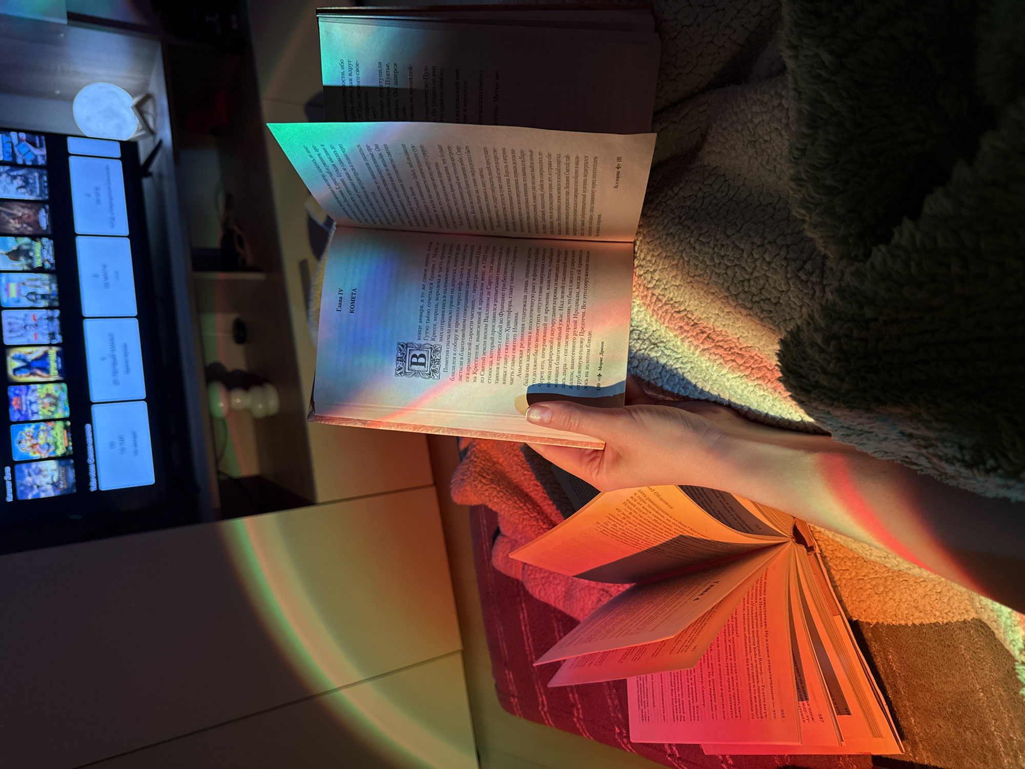 The image depicts a cozy, dimly lit indoor scene, likely at night, bathed in colorful light bands resembling a rainbow from a sunset lamp. A person, with their right hand visible, is holding an open book, actively engaged in reading. Another open book lies next to it, also open to a spread of pages. The setting features a textured, possibly sherpa-lined blanket or throw, suggesting a comfortable environment such as a bedroom or living room.
To the left, a television screen is on, displaying a grid interface typical of a streaming service, featuring various media thumbnails and some text in Cyrillic script, such as "Новые серии" (New series). A small, spherical, moon-shaped lamp rests on a surface near the television, emitting a soft glow.
The book held by the person is open to pages 190 and 191. Visible text includes "Глава IV" (Chapter IV), the title "КОМЕТА" (Comet), and the author's name "Морис Дрюон" (Maurice Druon) at the bottom of page 190. The text is in Cyrillic script.
The second open book, lying flat, shows pages with "Глава 4" (Chapter 4) and a title "След Оборотня" (Trace of the Werewolf). Other discernible Cyrillic text includes phrases like "Уважаемая доктор" (Dear Doctor) and "психиатр. Синклер" (psychiatrist. Sinclair), along with page numbers 314 and 355.
The colorful light from the lamp casts vibrant red, orange, yellow, green, and blue hues across the books, the person's hand, and the blanket, contributing to a warm and relaxed atmosphere.