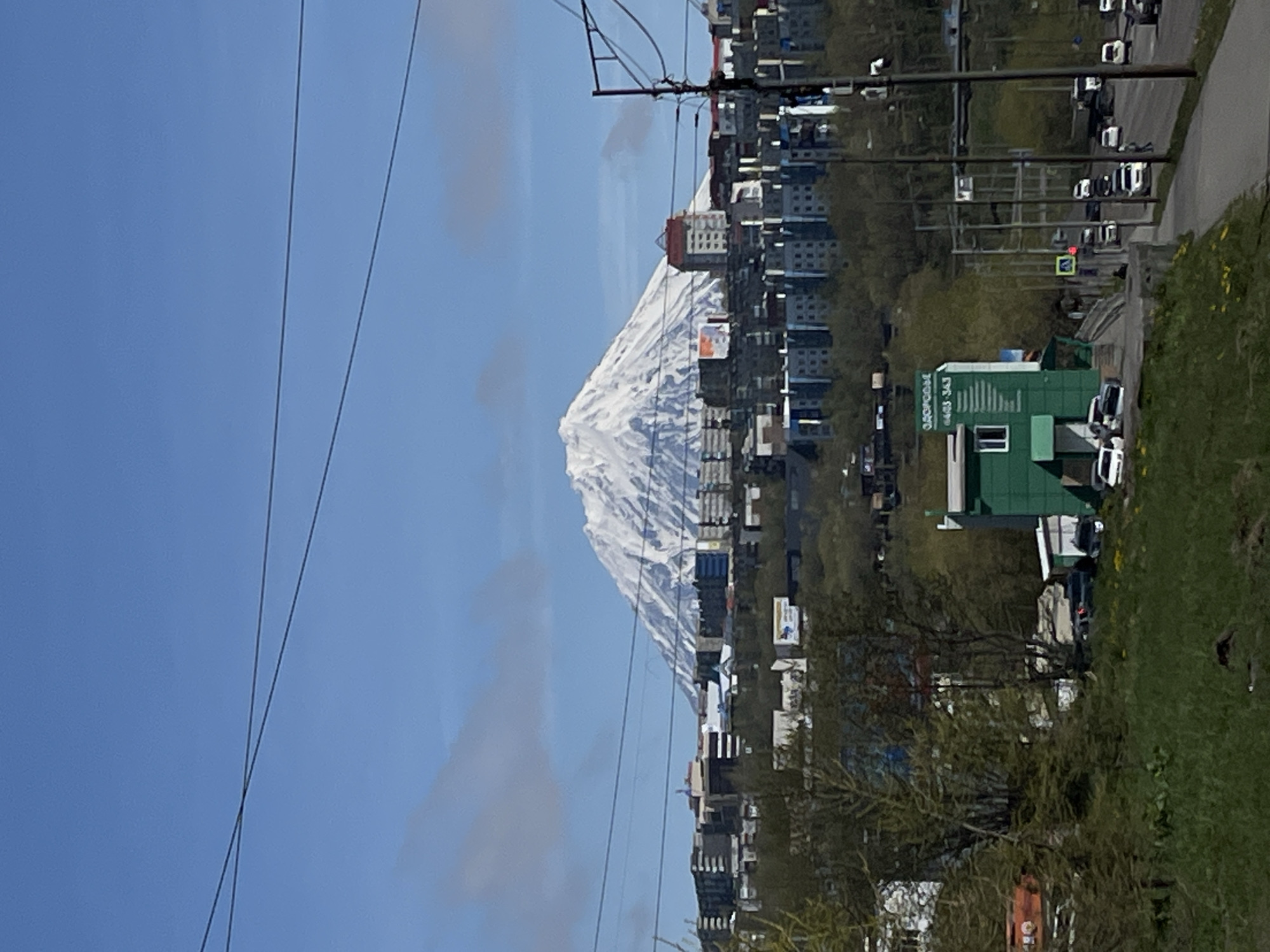The image shows a snow-covered mountain dominating the background, possibly a volcano. In the foreground, there's a city with buildings and urban infrastructure, including power lines and streets. Green vegetation, including grass and trees, is visible. A green building with a sign is prominent on the right side of the image. Cars are parked near the building. The location is likely Petropavlovsk-Kamchatsky, Russia, given the mountain's presence. The sky is blue with some clouds.