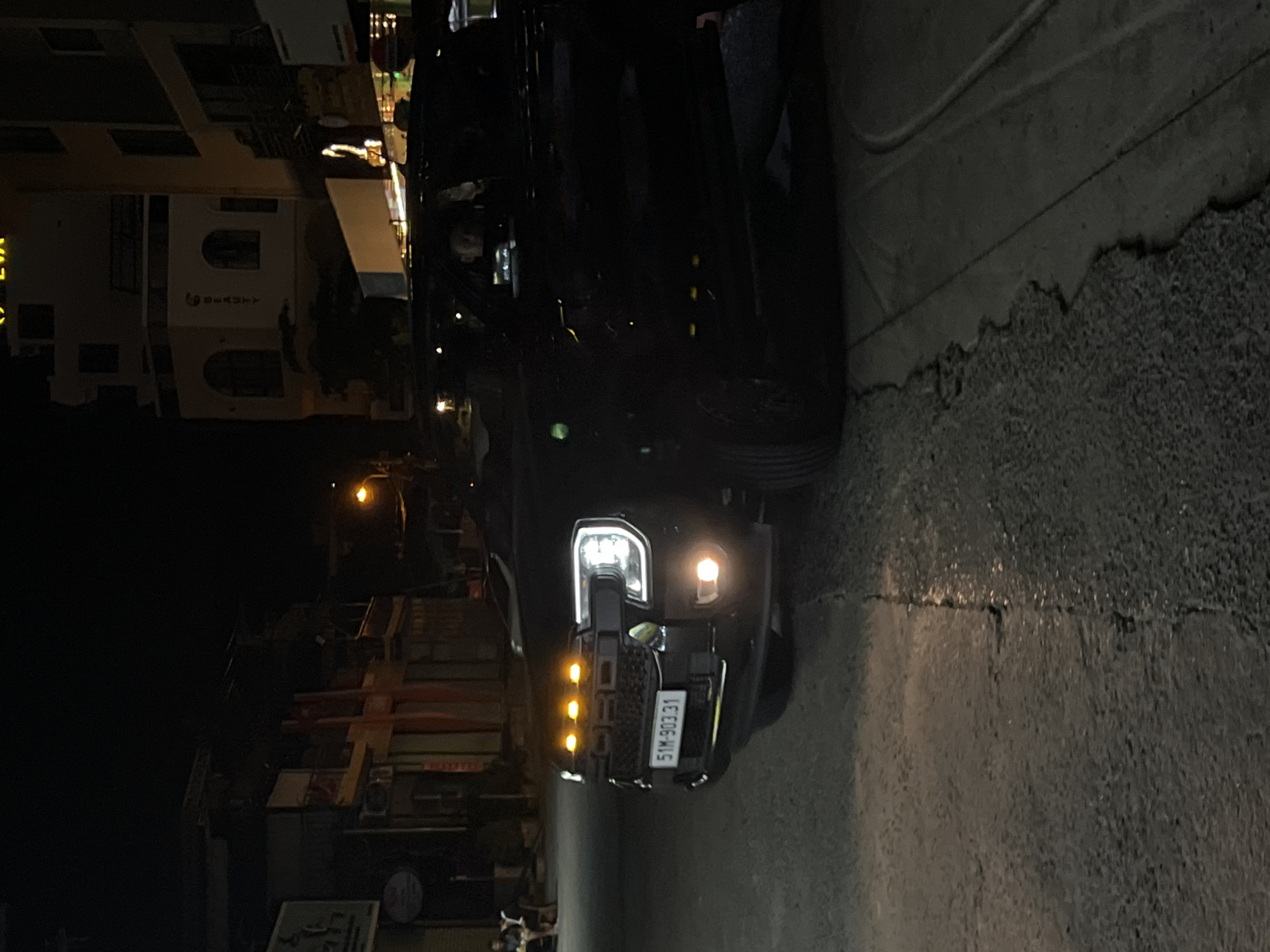 The image captures a dark street scene at night in Bac Nha Trang Ward, Vietnam, with a black pickup truck as the central subject. The truck's front is prominently displayed, featuring bright LED headlights and distinctive amber marker lights integrated into its grille. A white license plate, clearly visible on the front bumper, reads "51M-903.31".
To the left of the truck, several buildings line the street, partially illuminated by ambient light. One building reveals some architectural details and vertically oriented text that appears to be "SECURITY," though it is partially obscured and difficult to read entirely. The ground in the foreground is a dark, textured pavement or asphalt, showing some cracks and wear. A cable or power line runs across the upper right portion of the frame.
The overall scene is dimly lit, typical of nighttime, with artificial light sources, primarily from the truck, providing illumination. No people are clearly visible, and no specific activity is discernible beyond the truck being present on the street. The atmosphere is quiet and still.