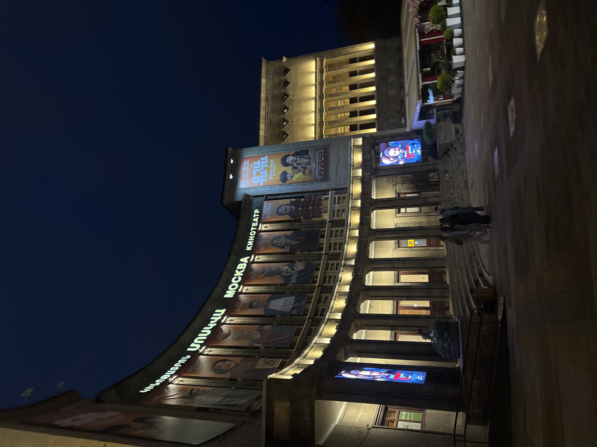 The image displays the Moscow Cinema (Մոսկվա Կինոթատրոն / Москва Кинотеатр) in Yerevan, Armenia, illuminated at night. The grand, multi-story building features classical architectural elements, including prominent columns and decorative lighting that highlights its facade against a dark blue sky.
The cinema's marquee curves along the building, displaying its name in bright green neon letters: "ՄՈՍԿՎԱ ԿԻՆՈԹԱՏՐՈՆ" (Moscow Cinema in Armenian) and "MOCKBA КИНОТЕАТР" (Moscow Cinema in Russian Cyrillic). Below this signage, numerous large movie posters are affixed to the building. One prominent poster features the title "TILO" along with the text "ԱՊՐԻԼԻ 29-ԻՑ" (From April 29th) in Armenian, depicting two men. Other posters show portraits and scenes related to various films. Several vibrant digital screens are also integrated into the building's exterior, showcasing movie advertisements.
In the foreground, a few individuals are walking on the pavement near a set of steps leading up to the cinema. To the right, an outdoor cafe or seating area with potted plants and white-striped barriers is partially visible, also lit by artificial lights. The upper sections of the building feature distinct window patterns and are bathed in warm light. The scene captures the lively ambiance of a city evening around a cultural landmark.