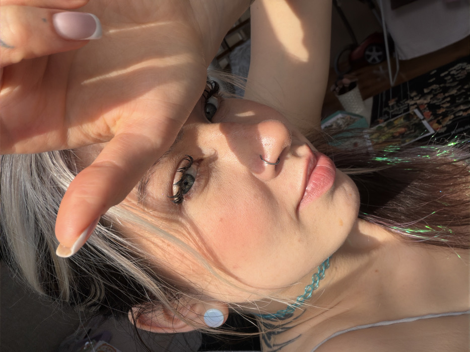 A close-up, sunlit portrait features a female individual, observed in Budva, Montenegro. The subject, appearing to be a young adult, possesses dark hair with distinct blonde/silver highlights and iridescent tinsel strands. Her right hand is raised, palm outwards, shielding her upper face from direct sunlight. Her left eye, light blue-grey with pronounced eyelashes, is open and directed towards the viewer. A silver hoop nose ring is present in her left nostril, and a light blue circular stud earring is in her left earlobe. Her lips are naturally colored, and her cheeks display a visible blush. She wears a light blue chain-link choker necklace, and a portion of a light-colored spaghetti strap top is visible on her left shoulder. A dark, indistinct tattoo is visible on her upper left back/shoulder area. The nails on her visible fingers exhibit a French tip manicure. The background is significantly blurred, consistent with an outdoor or semi-outdoor environment under strong sun exposure, and contains vague shapes indicative of distant structures or objects.