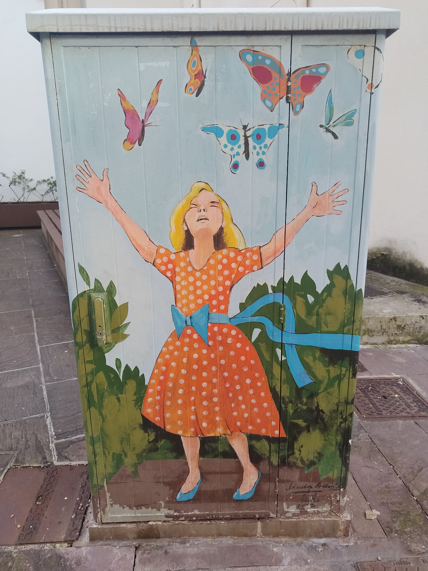 The image depicts a painted utility box located in Torrelavega, Spain. The painting features a girl with blond hair, wearing an orange dress with white polka dots and a blue bow, with her arms outstretched towards several butterflies flying above her. The lower portion of the box is painted with green foliage. The box is situated on a paved area with sewer grates visible in the ground nearby. The utility box is white and gray on the top. A signature in cursive is visible at the bottom right of the painted area.