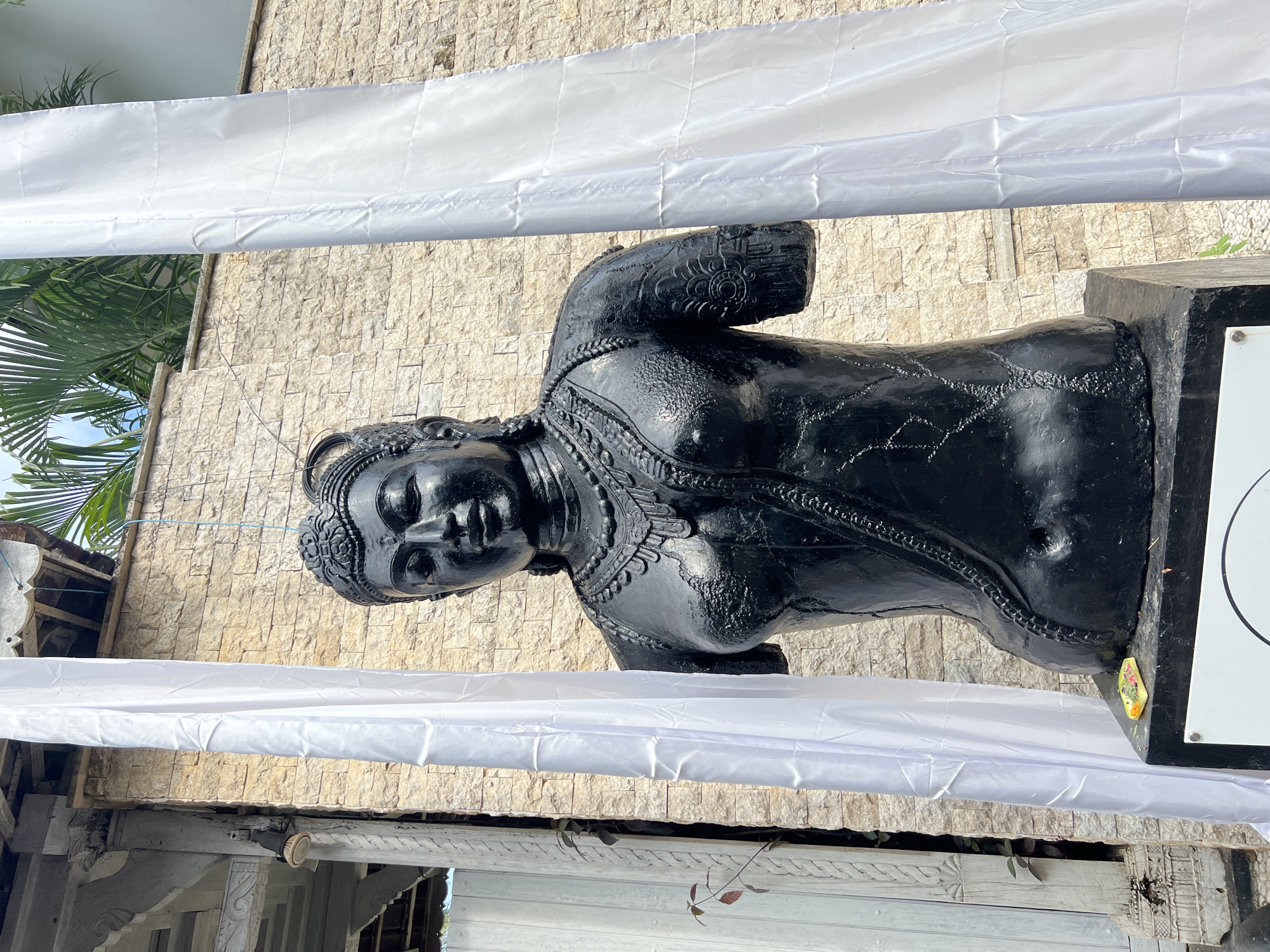 The image shows a black statue of a woman in Pecatu, Indonesia. The statue is positioned in front of a light-colored brick wall and partially obscured by two lengths of white fabric draped horizontally. The woman statue appears to be wearing some decorative clothing, and is mounted on a black pedestal. A tree or plant is also visible above the statue, and there is white decorated structure in the lower left of the frame.