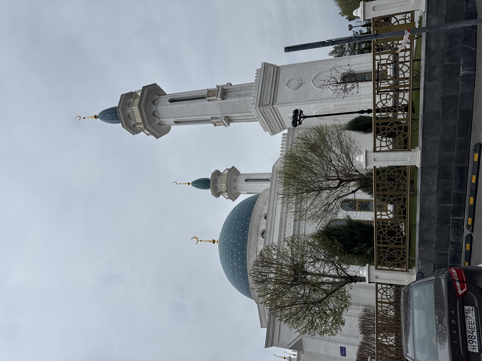 The image shows a mosque in Kazan, Russia. The mosque has a large blue dome with white stars, two white minarets with green tops, and a crescent moon at the top. In the lower right corner of the image, part of a black Lada car with a license plate is visible. The fence with patterned cutouts is on the right. Trees with leafless branches are in front of the mosque. The sky is gray and overcast.