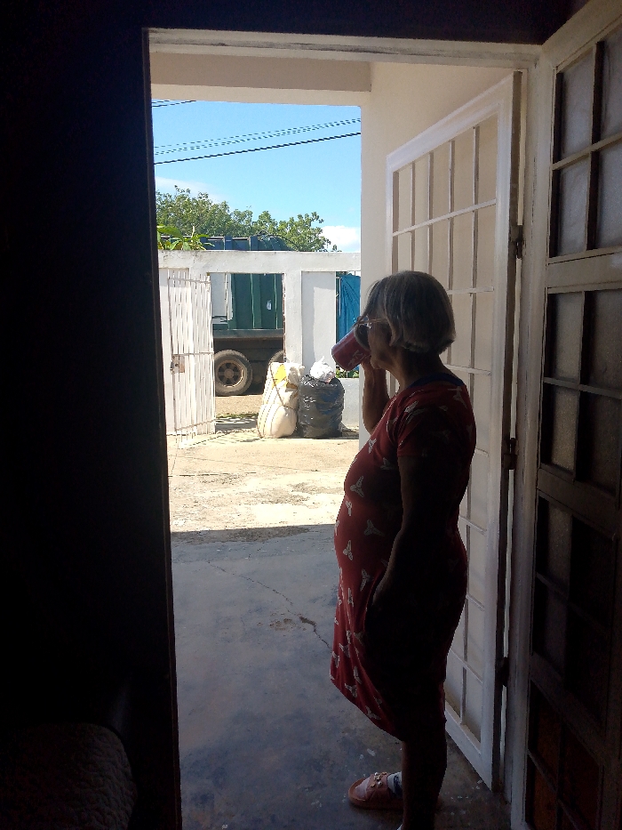 An older woman in a red dress and sandals drinks from a red cup, standing in the doorway of a white-framed house. A large, partially obscured truck is visible outside, parked near a white gate and some bags. The sky is blue with a few overhead wires. The view is partially obstructed by a dark, shadowy doorway in the foreground. The scene is sunlit.