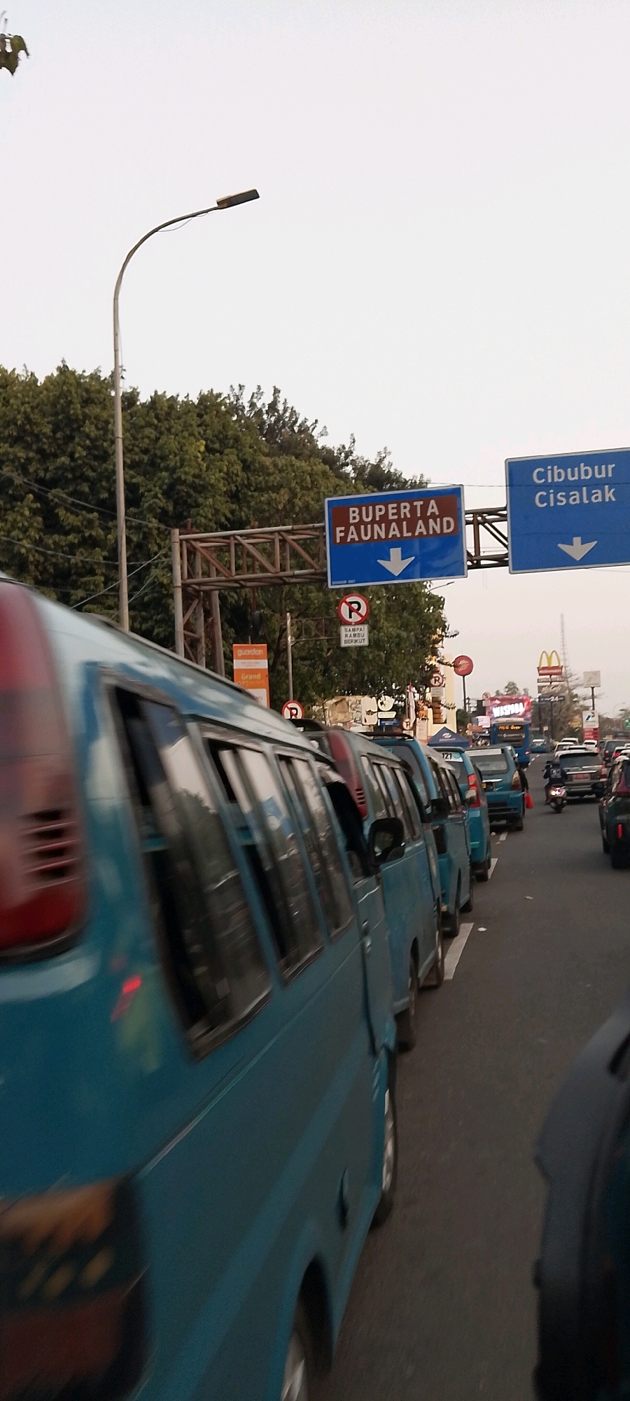 The image captures a traffic jam on a road in East Jakarta, Indonesia, likely during daytime. The foreground features the side of a blue minibus, suggesting it is in motion due to the motion blur. Several other blue minibuses are visible behind it, also in a line, indicating heavy traffic. Overhead, there are two blue road signs. The sign on the left reads "BUPERTA FAUNALAND" with a downward arrow, and the sign on the right reads "Cibubur Cisalak" with a downward arrow. There are also several red and white circular "P" signs with a diagonal line through them, indicating no parking zones, and other traffic signage. Trees line the side of the road, and a street lamp with a curved pole is visible in the upper left quadrant. In the distance, commercial signage for McDonald's is discernible, along with other vehicles, including motorcycles. The sky appears overcast.