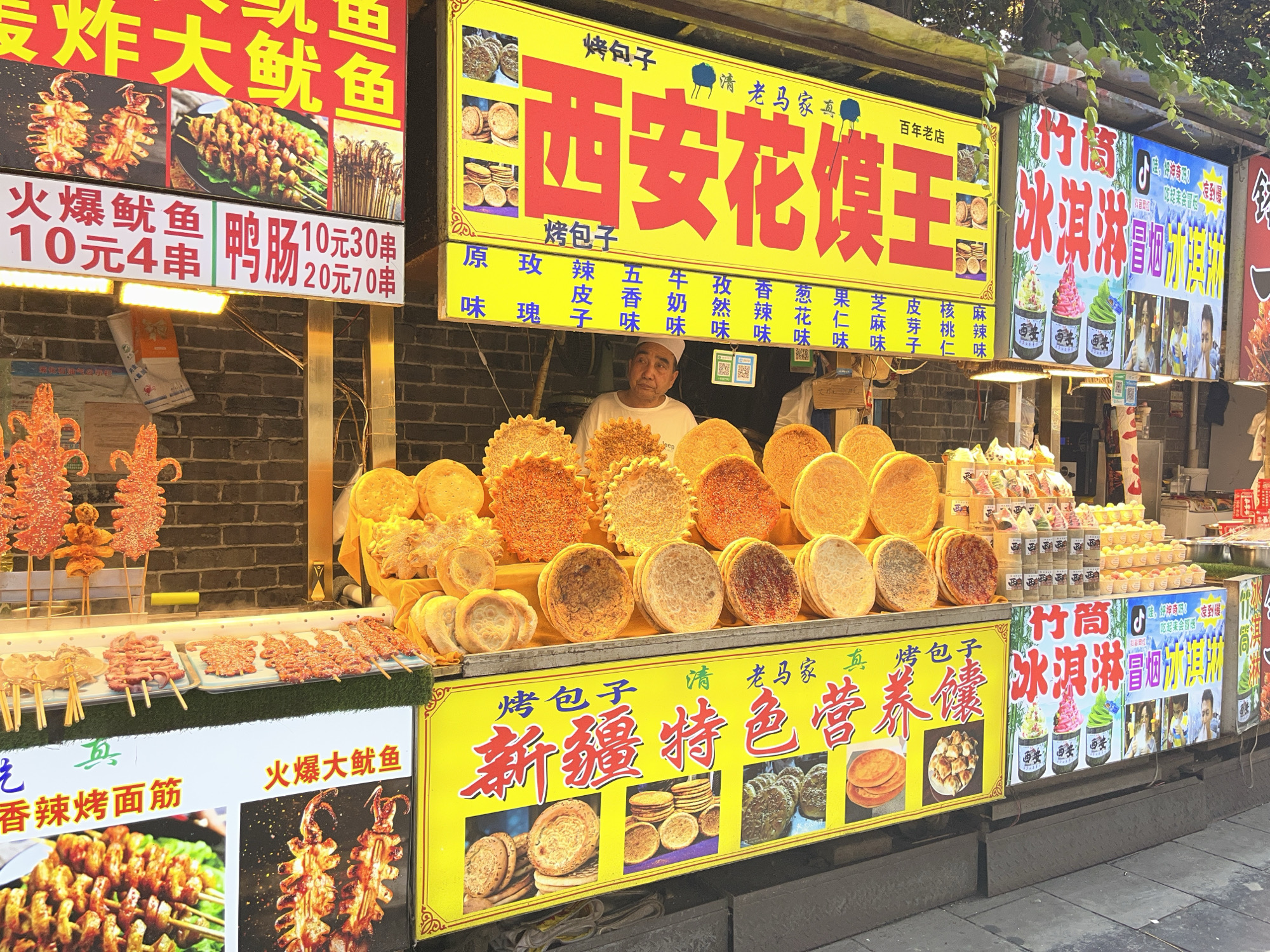 This is a street food stall in an outdoor market. The stall is selling various food items, including skewers of meat and seafood, flatbreads, and ice cream. The stall has several large signs with Chinese writing, advertising the food items. A man wearing a white hat and shirt is standing behind the counter, interacting with customers or preparing food. The stall is illuminated by bright lights, and there are green plants hanging overhead. The ground in front of the stall is paved with concrete. The scene appears to be in an urban environment, likely a busy street or market.