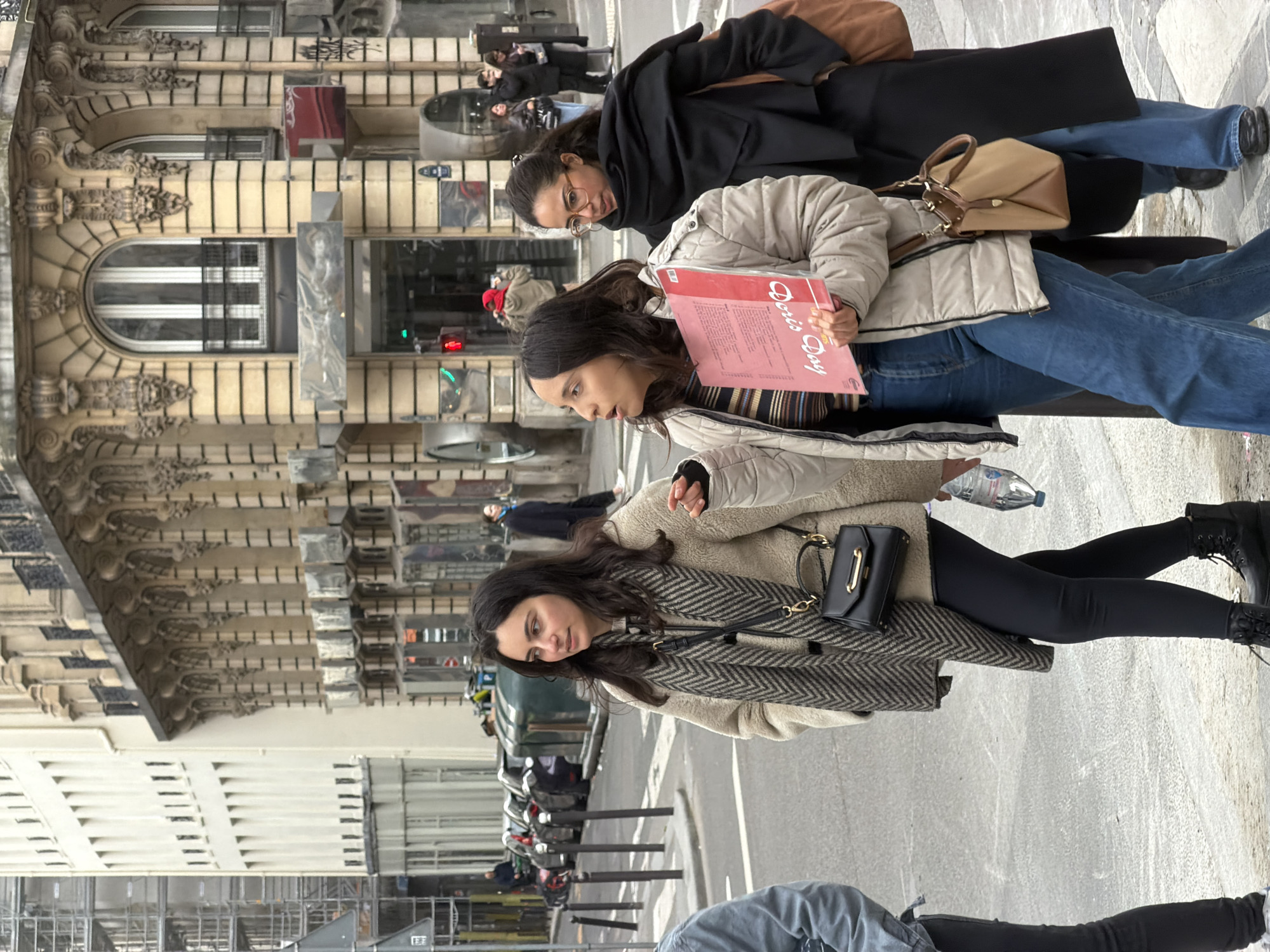 Three women are walking along a city street. One is holding a red album titled "Doris Day". They are dressed in casual clothing, including jeans and jackets. A tan building with ornate architectural details lines the street. There are parked scooters along the street. The location is most likely in Paris, France.