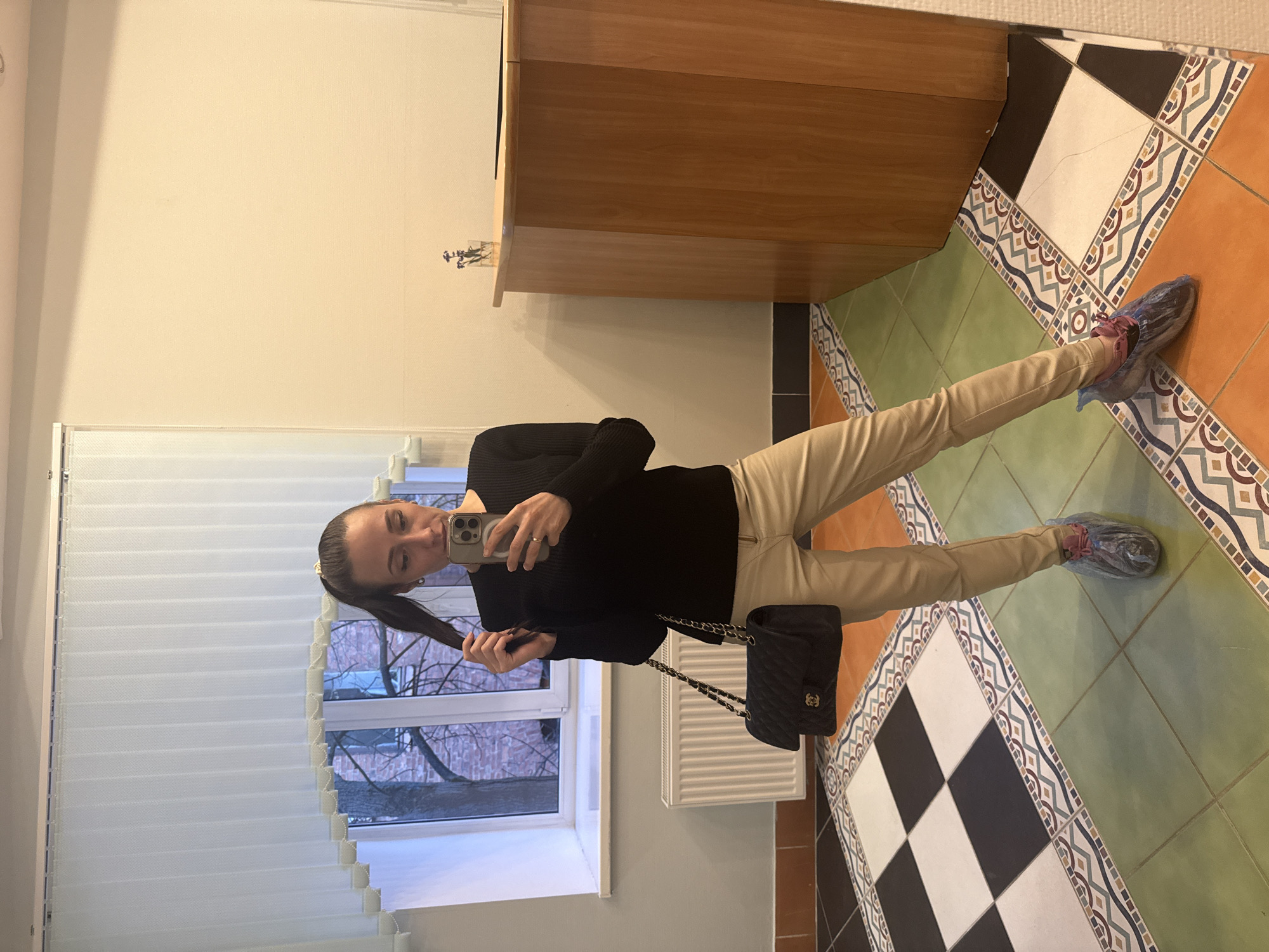 A woman is depicted indoors, taking a mirror selfie with a light-colored smartphone. She has dark hair styled in a high ponytail and wears a black ribbed long-sleeved sweater and light beige or khaki pants. On her left side, a black quilted Chanel shoulder bag with a gold chain strap is visible. Her feet, clad in dark sneakers with pink laces, are covered with blue disposable shoe covers. Her left hand is raised, seemingly adjusting or holding her ponytail.
The setting appears to be a room or hallway, potentially in Saint Petersburg, Russia. To the woman's left, a large window with white horizontal blinds is partially open, revealing bare tree branches and a red brick building outside. A white radiator is visible below the window. To her right, part of a light wooden cabinet or counter is in the frame. The floor is distinctive, covered in a mosaic of ceramic tiles featuring large green and orange squares, geometric patterned borders in blue, red, and white, and sections of black and white checkerboard tiles.
The activity is a self-portrait being taken in a mirror. The time of day appears to be daytime, with natural light entering from the window, augmented by indoor lighting. The bare tree branches outside suggest late autumn or winter weather. The disposable shoe covers are a notable detail, common in Russia for maintaining cleanliness in various indoor environments like homes, clinics, or museums. No specific text is clearly readable, except for the Chanel logo on the handbag.