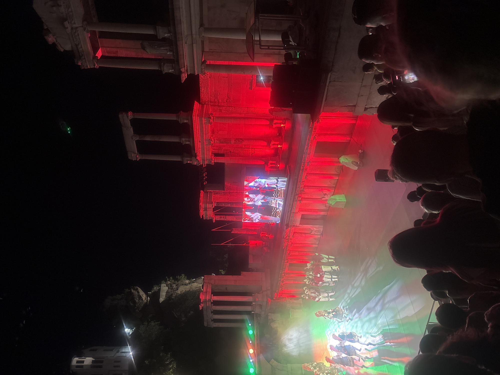 The image depicts an outdoor performance at the Ancient Roman Theatre in Plovdiv, Bulgaria. The theatre is illuminated with vibrant red and green lights, casting a dramatic glow on the ancient stone structure. On the stage, performers in traditional costumes are engaged in a dance or theatrical presentation. A large screen displays images of performers, likely part of the show. The audience is visible in the foreground, silhouetted against the stage lights, suggesting the event is taking place at night. The architectural grandeur of the Roman theatre is evident, with its tiered seating and imposing stone columns. The scene captures a cultural event in a historic setting, highlighting the juxtaposition of ancient architecture and modern performance.