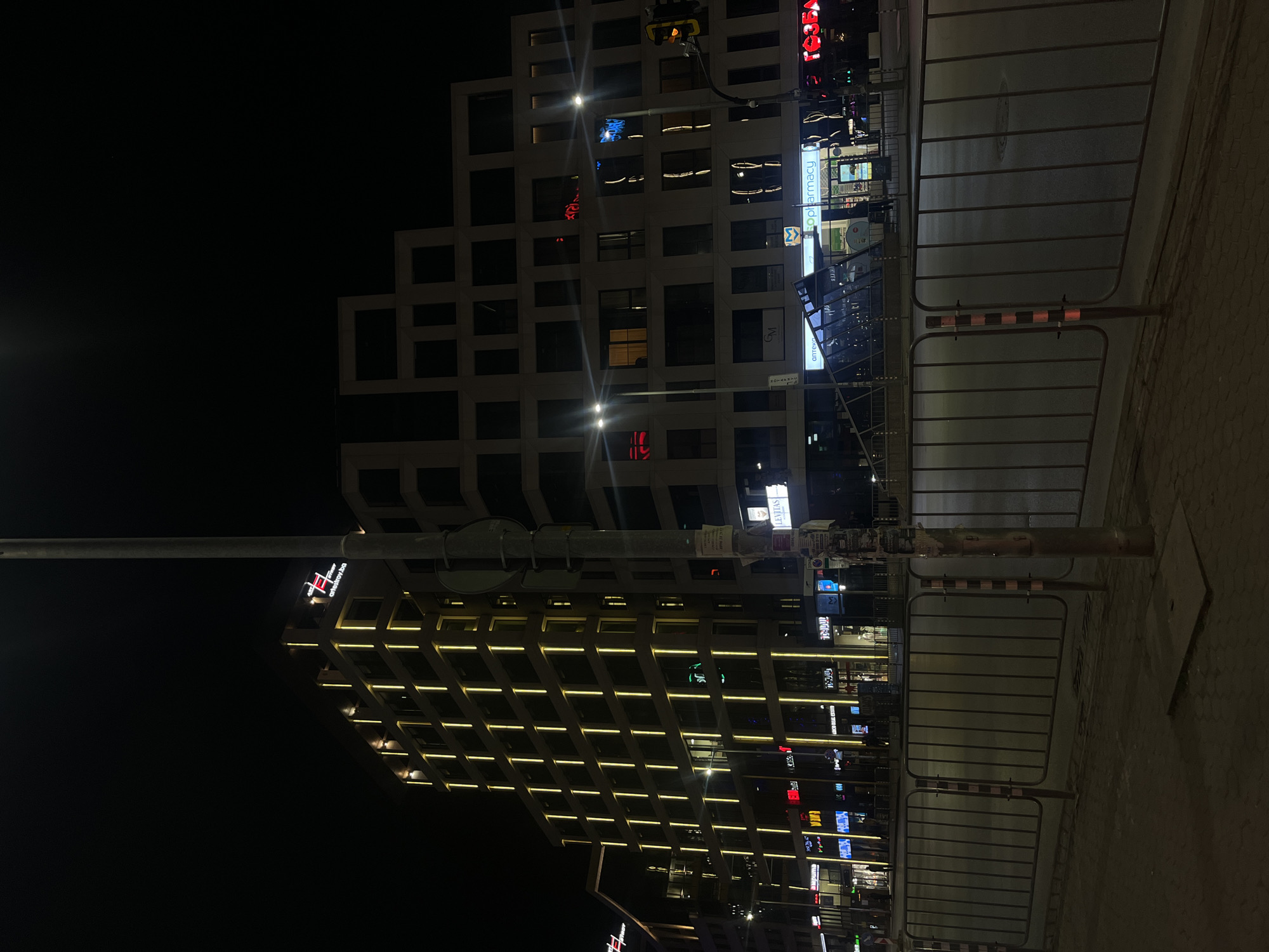 The image depicts a nighttime urban scene in Sofia, Bulgaria. Two modern buildings dominate the view, each with a distinct architectural style. One building features a staggered, geometric facade with lit windows, while the other is characterized by horizontal lines of light accentuating its structure. Streetlights and neon signs illuminate the scene. Pedestrian barriers are visible in the foreground. The time of day is evening or night. No people are seen in the image.