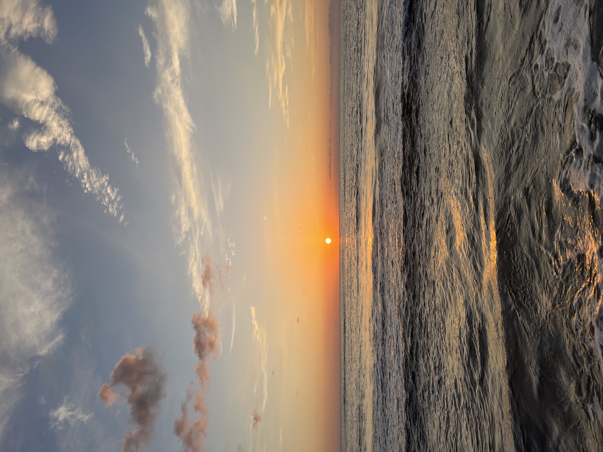 The image captures a serene sunset over the sea. The sun, a bright orange orb, is low on the horizon, casting a warm glow across the sky and the water. Wispy clouds, tinged with pink and orange, are scattered across the blue expanse. The sea below is calm, with gentle waves rolling towards the shore, reflecting the vibrant colors of the sunset. The foam of the breaking waves creates a soft contrast against the darker water. While the image evokes a sense of tranquility and natural beauty, there is no information available to suggest it is specifically located in Isola delle Femmine, Italy. The scene is devoid of any people or specific objects that would indicate a particular location within a city.