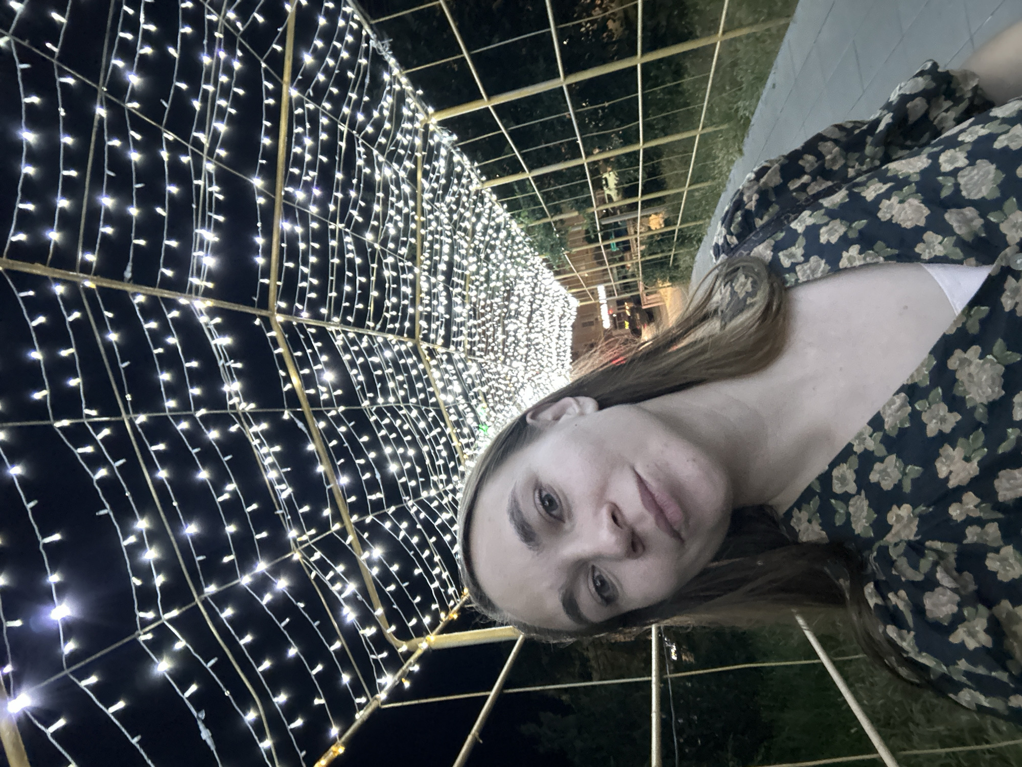 A young woman with long brown hair is taking a selfie in a pedestrian tunnel adorned with white string lights. She is wearing a dark dress with a floral pattern. The tunnel appears to be outdoors, with trees and a street visible in the background. The image is likely taken at night, given the artificial lighting. The location is the city of Elista, Russia.