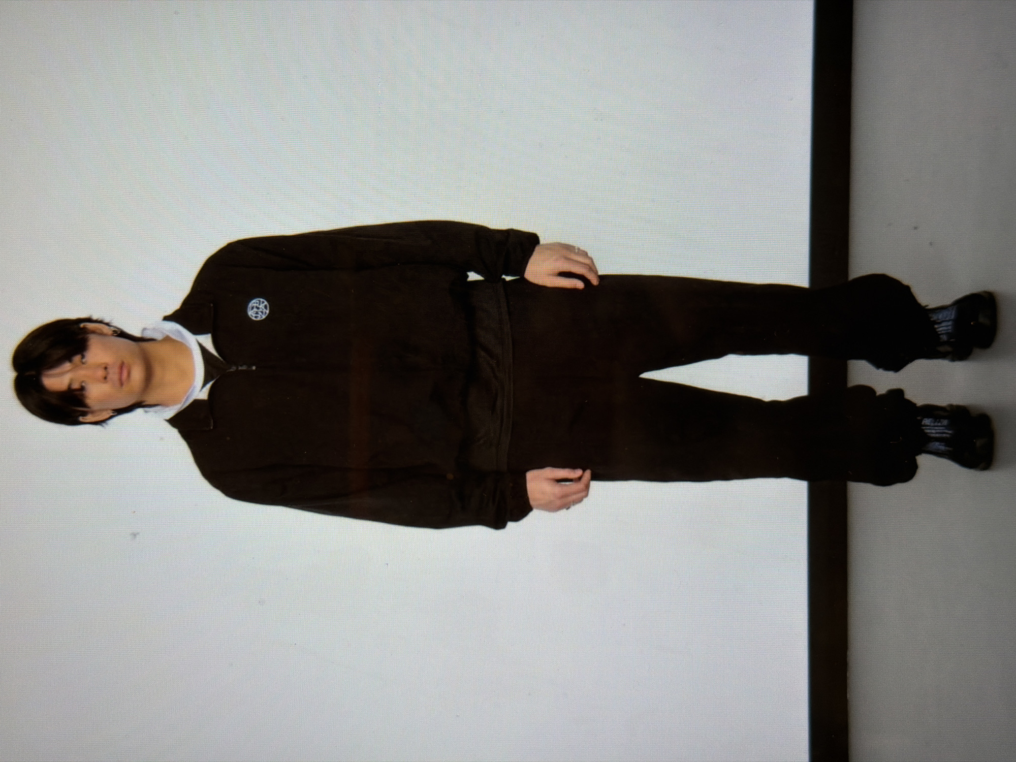 A person wearing a dark-colored tracksuit stands against a white wall. The person has dark hair and is wearing black shoes. A dark, vertical line appears on the right side of the image. The location is not discernable.