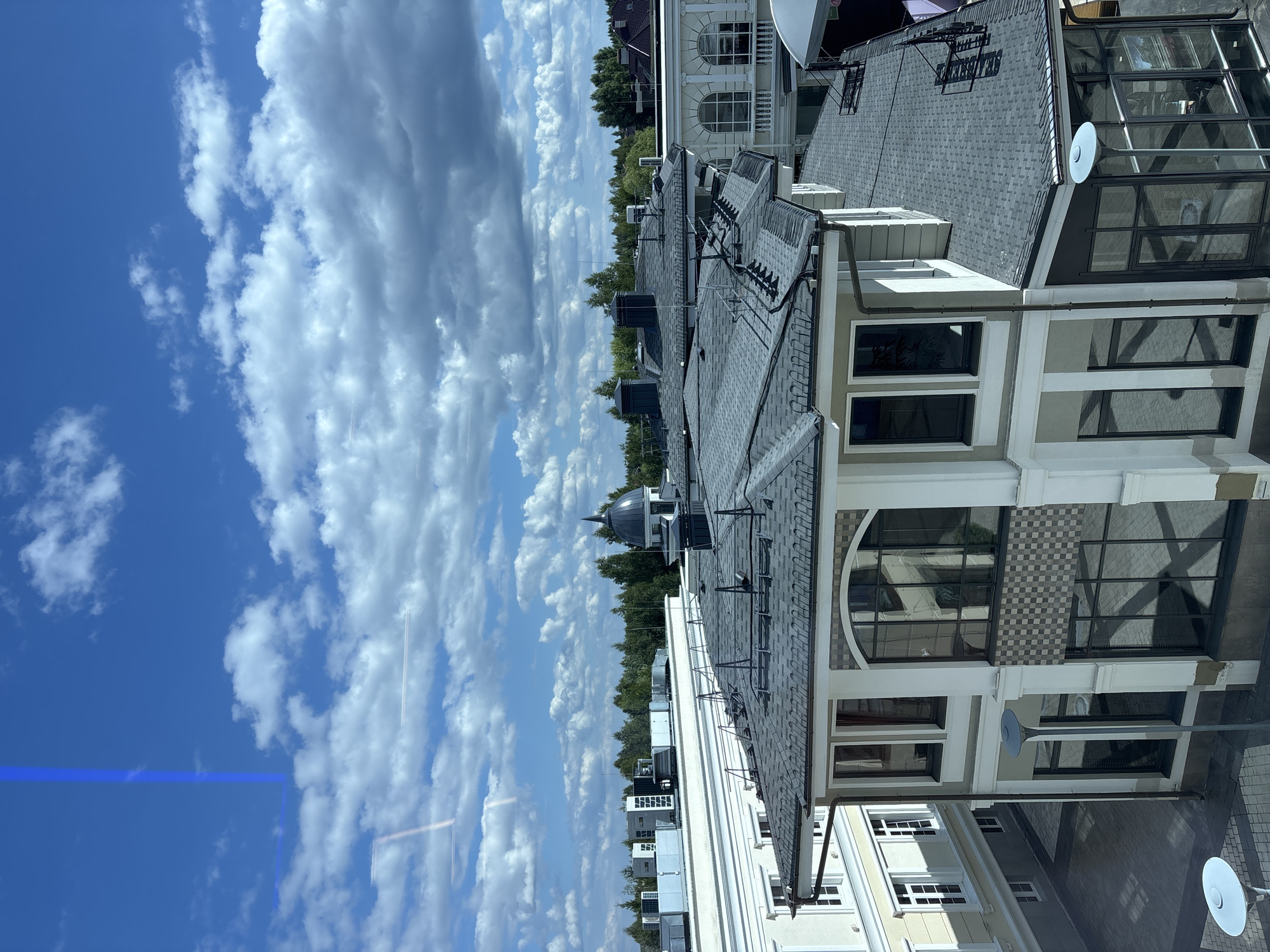 This is a high-angle, medium shot of a modern suburban area under a partly cloudy blue sky. The foreground features a multi-story building with a dark gray shingle roof and a light-colored facade. The building has numerous large windows, some of which reflect the sky and surrounding architecture. There are also visible air conditioning units on a distant rooftop, and a dark dome structure with a spire. The scene is captured during the daytime, with shadows indicating the sun is to the left. The location is identified as Monolit, Russia.