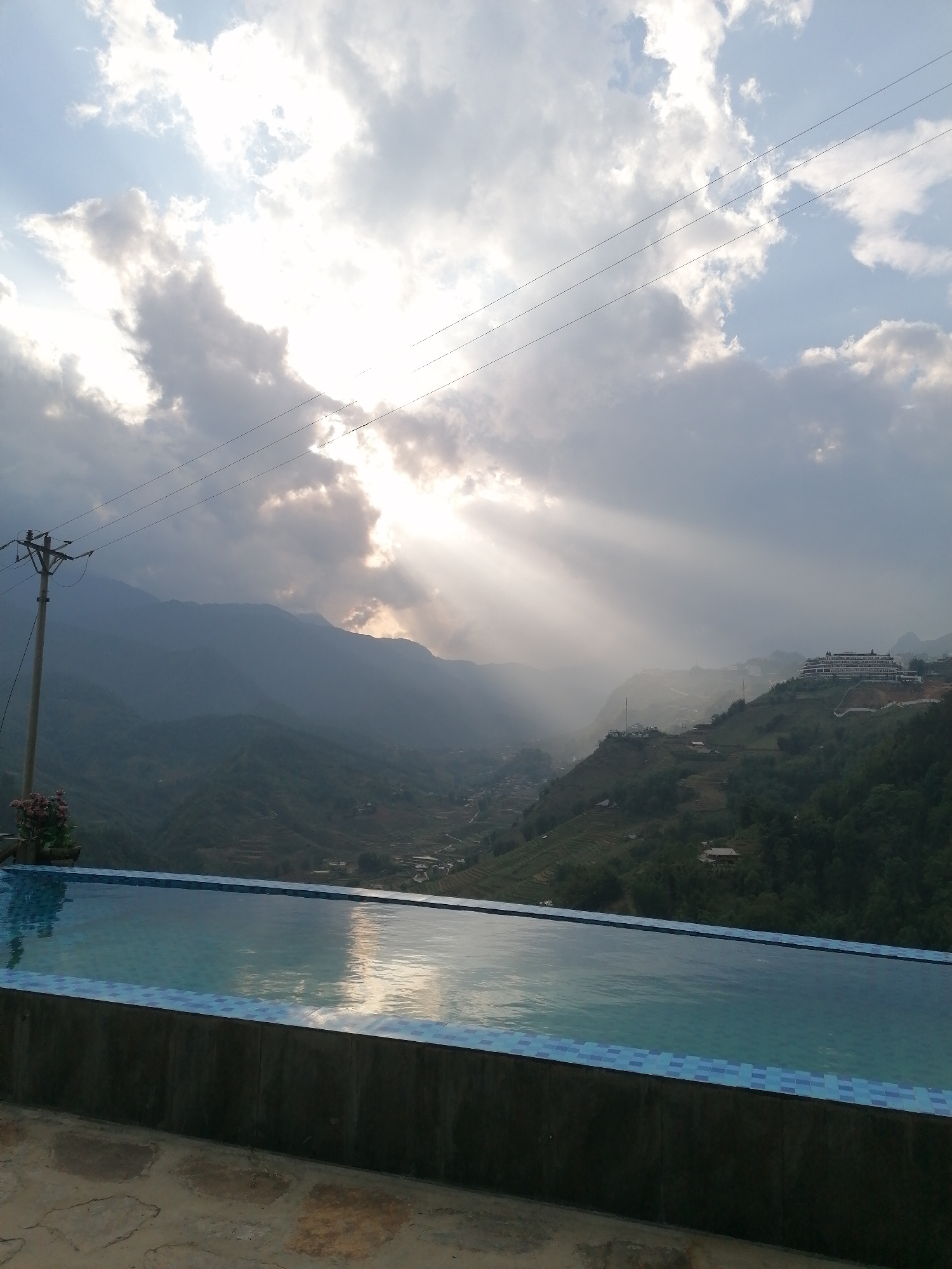 This media file captures a majestic panoramic view of a mountainous landscape in Sa Pa Ward, Vietnam, likely taken from a high vantage point overlooking a valley. In the foreground, an infinity pool with blue and white mosaic tiles lines the edge, reflecting the sky and sun. The water in the pool is calm, with subtle ripples. Below the pool, a rough stone patio is visible.
Beyond the pool, the midground features a vast expanse of verdant mountains and valleys, with terraced fields visible on some slopes, indicative of agricultural activity. Scattered buildings and small villages nestle within the valleys and cling to the hillsides. On a prominent ridge in the distance, a large, modern resort-like building with multiple levels can be seen. A utility pole with power lines runs across the left side of the frame, extending into the distant sky.
The sky dominates the upper half of the image, filled with dramatic, partially cloudy conditions. Rays of sunlight, often called crepuscular rays, spectacularly break through a gap in the clouds, illuminating the valley and creating a striking visual effect. The sun appears to be low in the sky, suggesting either an early morning sunrise or a late afternoon sunset. The overall atmosphere is serene and awe-inspiring, highlighting the natural beauty of the mountainous region. There are no people visible in the image, and no discernible text.