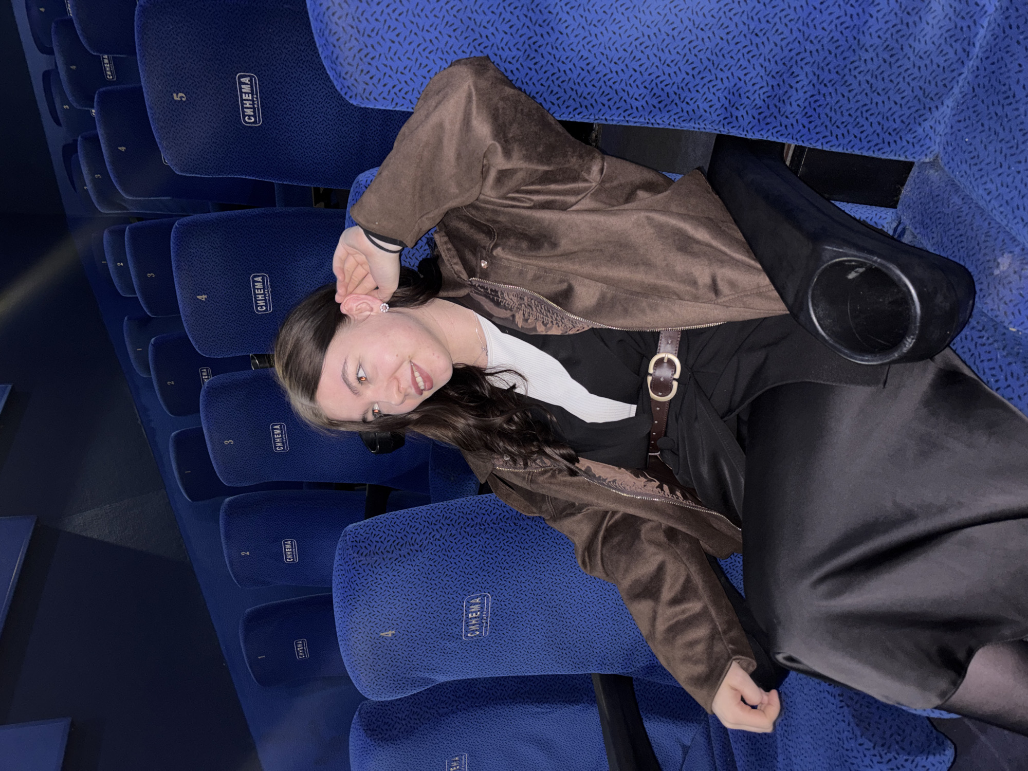 A young woman sits diagonally in a blue patterned movie theater seat with the word "СИНЕМА" (Cinema) and a number printed on the headrest. She wears a brown jacket, a black shirt, a white undershirt, and black pants with a brown belt. The theater appears mostly empty. The location is potentially in Perm, Russia based on the Russian word on the seats.