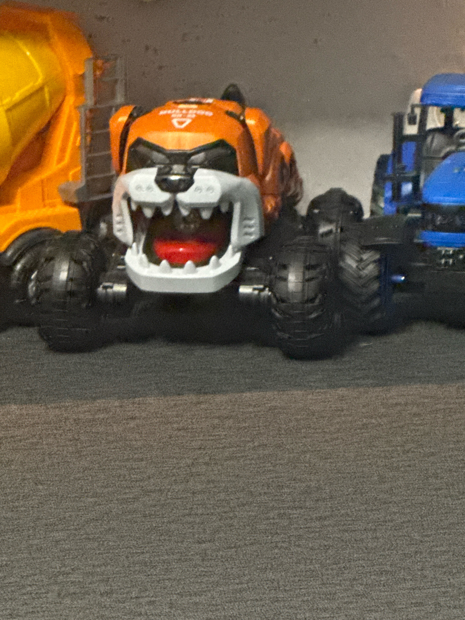 The image shows a close-up of toy vehicles. In the foreground, a toy truck with a tiger-like face is prominent. This truck is orange and white with a large, open mouth displaying sharp teeth and a red tongue. It has large black wheels. To the left of the tiger truck, a portion of a yellow toy cement mixer truck is visible. To the right of the tiger truck, a blue toy truck is partially visible. The toys are placed on a grey, textured surface, possibly a carpet or rug. The background is a muted, grey wall. There is no indication of a specific location within the city of Gorzow, Poland, or any people present in the image.