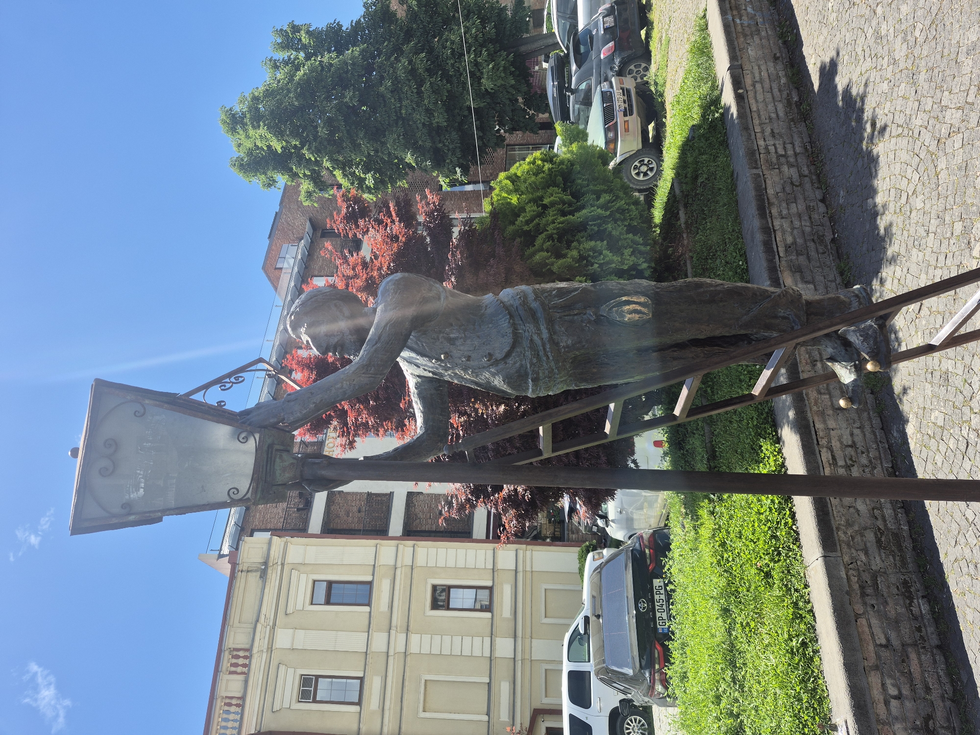 The image captures a statue of a man positioned on a ladder, reaching towards a streetlight. The statue appears to be made of bronze or a similar material, given its dark, weathered texture. The streetlight is mounted on an ornate metal frame, which has intricate designs.
Behind the statue and to the right, there is a row of parked cars and SUVs. These vehicles are diverse in make and color. A green tree is visible in the background, along with sections of buildings. One building is a light cream color with noticeable windows.
The background includes trees with red leaves and other foliage, suggesting an urban environment. The pavement is laid with cobblestones. The location is the city of Tbilisi, Georgia.