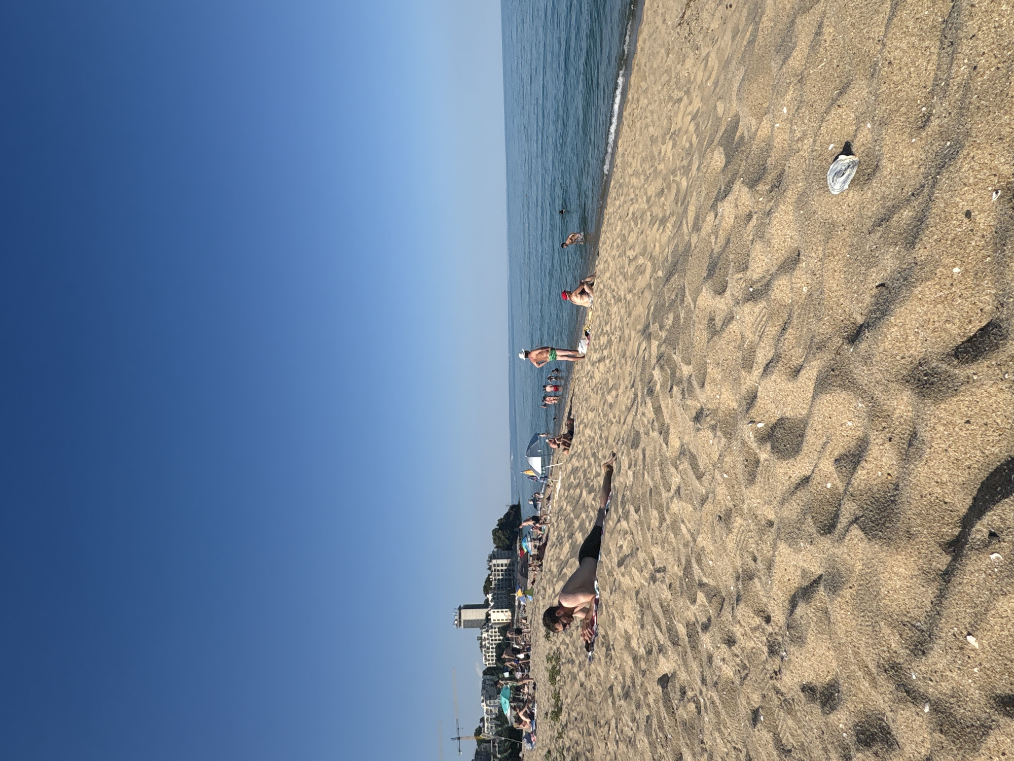 A sunny day at a crowded beach is depicted from a low-angle perspective, with the clear blue sky taking up the left two-thirds of the frame. The ocean, a tranquil blue, meets the sandy shore on the right. The beach is populated with numerous people, many of whom are in the water or relaxing on the sand. Umbrellas dot the shoreline, offering shade to beachgoers. In the background, a strip of buildings with white facades and balconies suggests a coastal town. A construction crane is visible to the far left. The sand itself features ripples from the tide or wind, with a few scattered shells and small white pebbles. A single person is prominently featured in the foreground, lying on their stomach on the sand, appearing to be sunbathing or resting. The scene evokes a feeling of leisure and warm weather. No text is visible in the image.