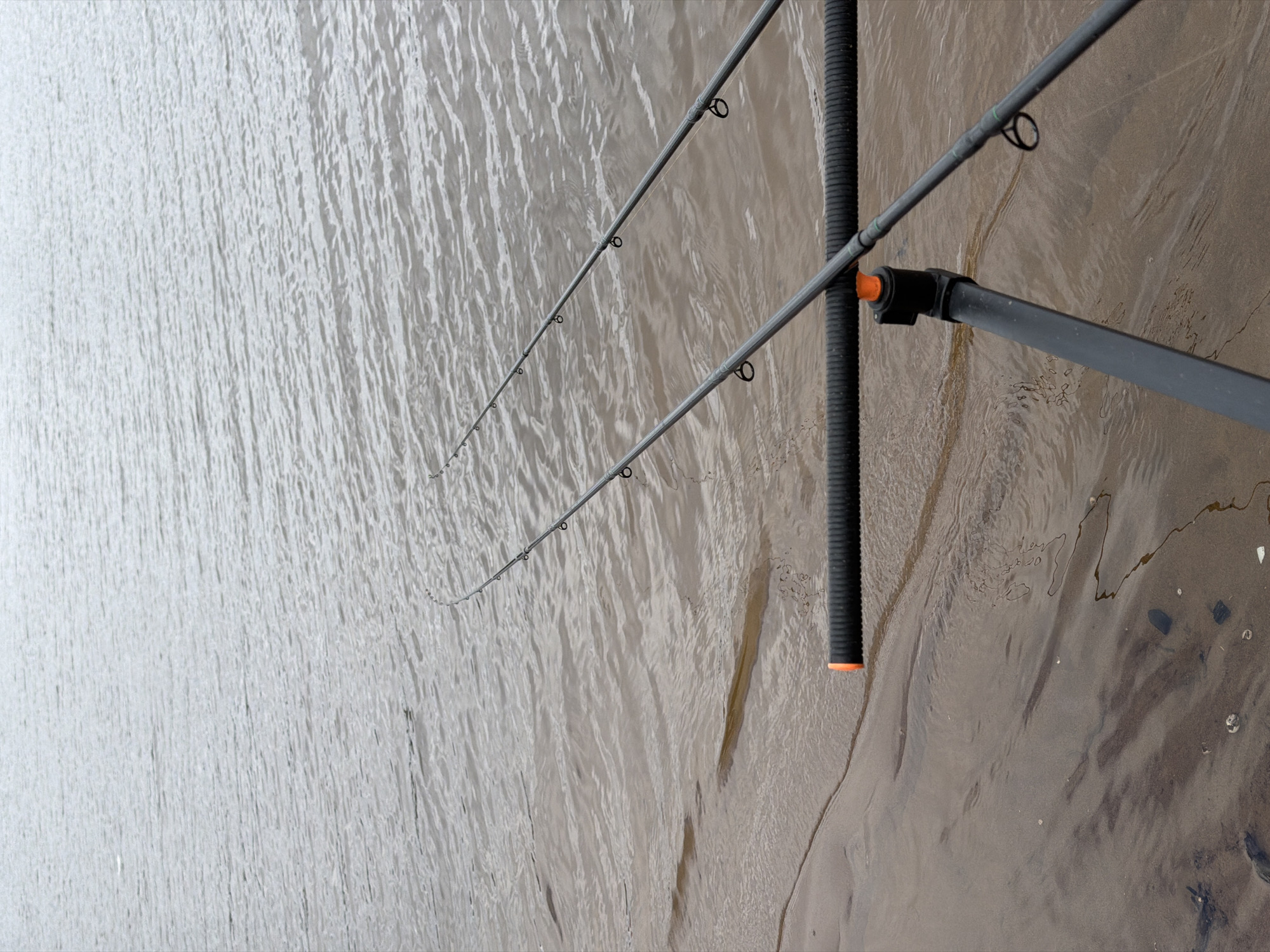 The image depicts a fishing scene, likely set on a riverbank in Chaikovsky, Russia. Two fishing rods are extended over a body of water, suggesting an active fishing session. The rods are supported by a black, corrugated rod rest, adding stability. The water appears murky, hinting at sediment or recent disturbance. The surrounding environment is partly visible, revealing what seems to be a muddy bank or shoreline. The scene is static, with no explicit human presence evident beyond the implied activity of fishing. The composition suggests a focus on the fishing activity itself, with the environment providing context.