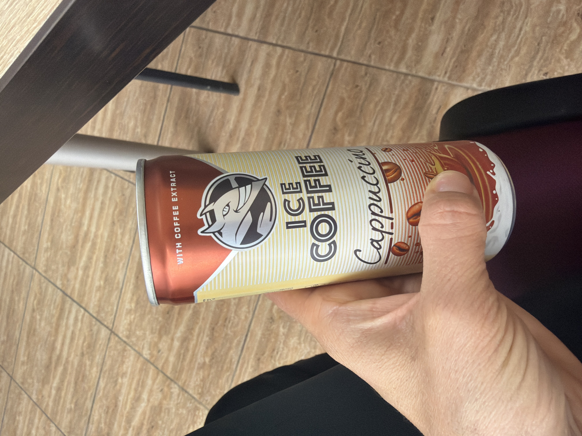 A can of Ice Coffee Cappuccino is being held by a person's hand. The can is cream-colored with vertical lines, and the words "ICE COFFEE Cappuccino" are printed in black. A brown and white logo is visible on the can, and there are coffee bean illustrations near the top. The person is wearing black pants or a skirt. A table leg and part of the floor, which is covered in a tile pattern, are also visible. The photo's location cannot be determined from the image.