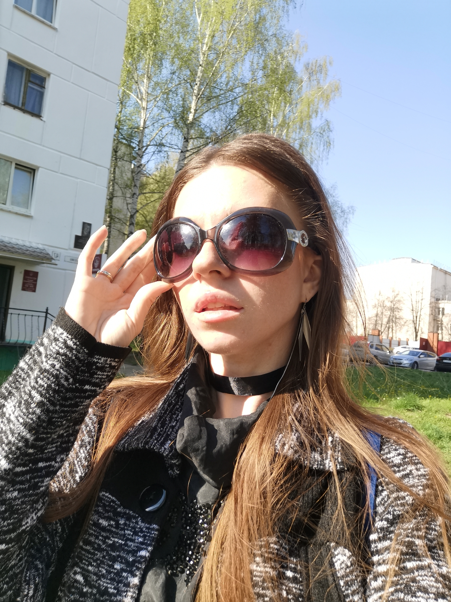 A fair-skinned woman with long, light brown hair is the central subject of the photograph. She is wearing large, round sunglasses with dark frames and gradient lenses. The woman is dressed in a black and white knit cardigan over a black top, and she has a black ribbon choker around her neck. She is wearing a ring on her left hand. Her right hand is slightly raised, adjusting or holding her sunglasses. Her expression appears neutral.
The background includes a white building with a green door and window frames. In the distance, there is a glimpse of other buildings and cars. Bare trees with some green foliage suggest it is either early spring or late fall. The setting appears to be an urban area, with a patch of green grass visible in the lower right corner. The lighting indicates it is a sunny day.
The location is in a city of Unknown, Unknown.