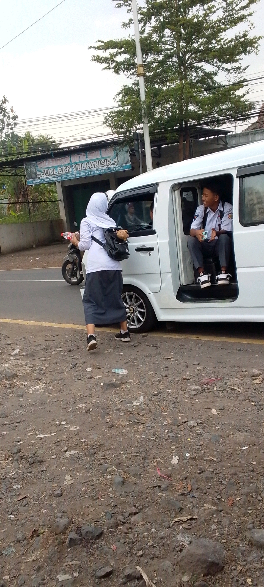 A young woman wearing a white hijab and a dark gray skirt stands on a gravelly roadside in Cilaku, Indonesia. She is holding a red bottle and a small object, possibly a toy, is attached to her black backpack, which has a teddy bear hanging from it. Beside her, a white van is parked with its sliding door open. A young man in a school uniform, consisting of a white shirt and dark pants, sits inside the van, looking towards the woman. A motorcycle is visible to the left of the scene. In the background, a banner reads "JUAL BAN VULKANISIR" and contains text related to locations and phone numbers in the Cilaku and Cianjur areas. The environment appears to be an overcast day, with trees and power lines visible overhead.