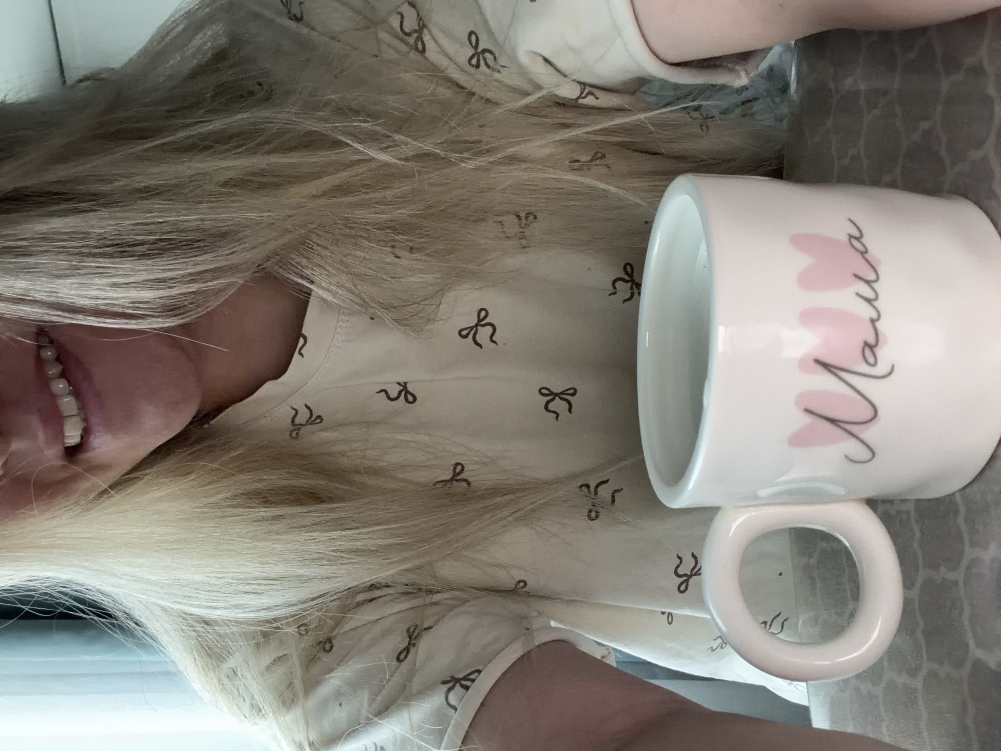 The image, captured from a high angle and rotated 90 degrees clockwise, presents a close-up of a person holding a mug in an indoor setting in Votkinsk, Russia. The primary subject is a person, whose face is partially visible from the nose down, revealing a wide, joyful smile with bright white teeth. Long, flowing blonde hair, appearing light and possibly streaked, dominates a significant portion of the frame, fanned across the person's shoulder and chest.
The person is wearing a light-colored garment, likely a casual top or sleepwear, which is patterned with numerous small, repeating dark brown or black bow-tie or ribbon designs. A light-skinned arm is also visible towards the bottom right, suggesting the person is holding the mug.
A prominent object in the image is a white ceramic mug with a distinctive, chunky circular handle. The mug is adorned with two pink heart shapes and the word "Мама" (Mama) written in elegant, dark cursive script. The background features a patterned surface, possibly a tablecloth or countertop, with a muted grey, abstract, or geometric design. The overall scene conveys a sense of warmth and contentment, highlighted by the person's happy expression.