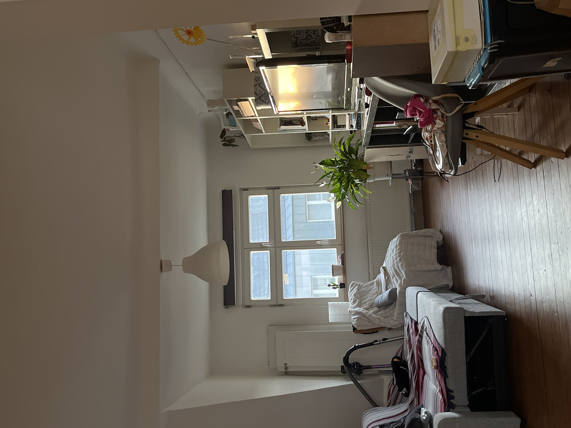 The image shows an interior living space in a residential building. The room is well-lit with natural light coming through a multi-pane window. A ceiling lamp with a simple, white lampshade hangs from the ceiling. A white radiator is under the window.
The room is furnished with a chair covered with a blanket, and a gray couch with striped throw blankets.
Along the opposite wall, there is a shelf or storage unit filled with various items, including books and decorative objects. There is a green plant on the shelf. A workstation with a large monitor, stands on a wooden frame, also sits here.
The room appears to be in Saarbrücken, Germany, based on the style of architecture visible through the window. No people are visible in the image. The environment suggests a comfortable and functional living space.