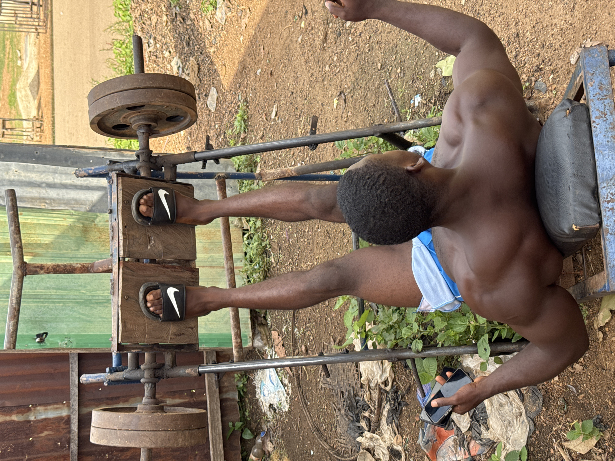 A dark-skinned man is exercising on a makeshift leg press machine outdoors. The machine appears to be constructed from scrap metal and wood, with weight plates attached to metal rods. The man is wearing blue and white shorts and black Nike slides. He is holding a smartphone in his left hand. The background shows a dirt ground with some scattered debris and vegetation, suggesting an outdoor location in a rural or semi-urban area. The general setting is consistent with an informal outdoor gym. The image appears to have been taken in Akwanga, Nigeria.