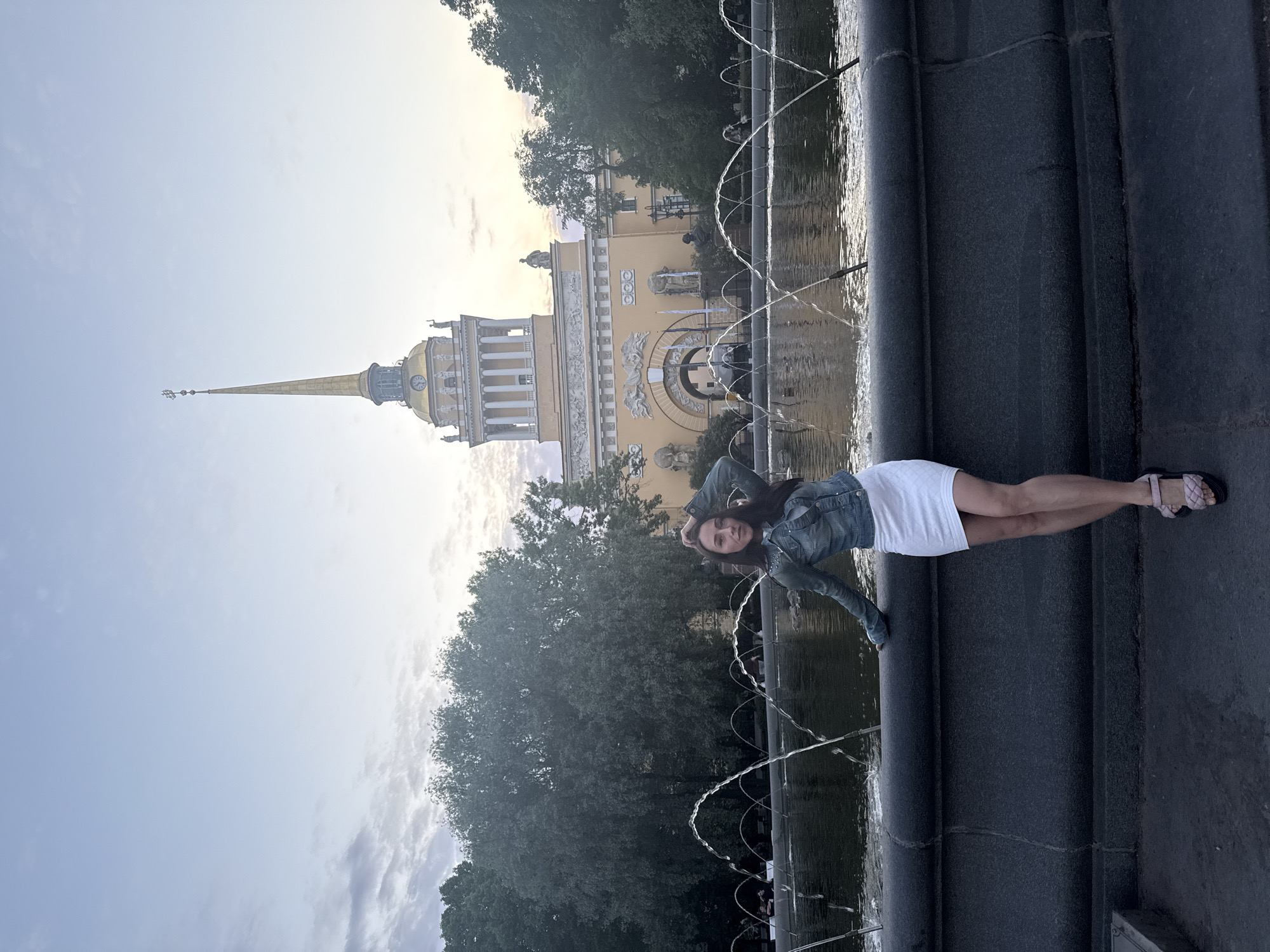 An outdoor, horizontally oriented photograph depicts a young woman posing in front of a grand architectural landmark and a fountain in Saint Petersburg, Russia. The woman, with long dark hair, wears a light blue denim jacket over a white skirt or dress and light-colored sandals. She stands casually with one leg slightly bent, looking towards the camera with a gentle smile.
In the background, a majestic, light yellow building with classical columns and a prominent golden spire, characteristic of the Admiralty Building, stands under a bright, partly cloudy sky. The building features intricate facade details and a clock visible on its tower. Green foliage and trees are visible on either side of the building and behind the fountain, adding natural elements to the urban scene.
In the foreground, a dark stone fountain edge runs across the frame, with multiple arcing jets of water creating a dynamic visual element. The light suggests it is late afternoon or early evening, casting a soft glow on the scene. The overall impression is one of a relaxed moment captured in a historically significant and picturesque location. No visible text is present in the image.