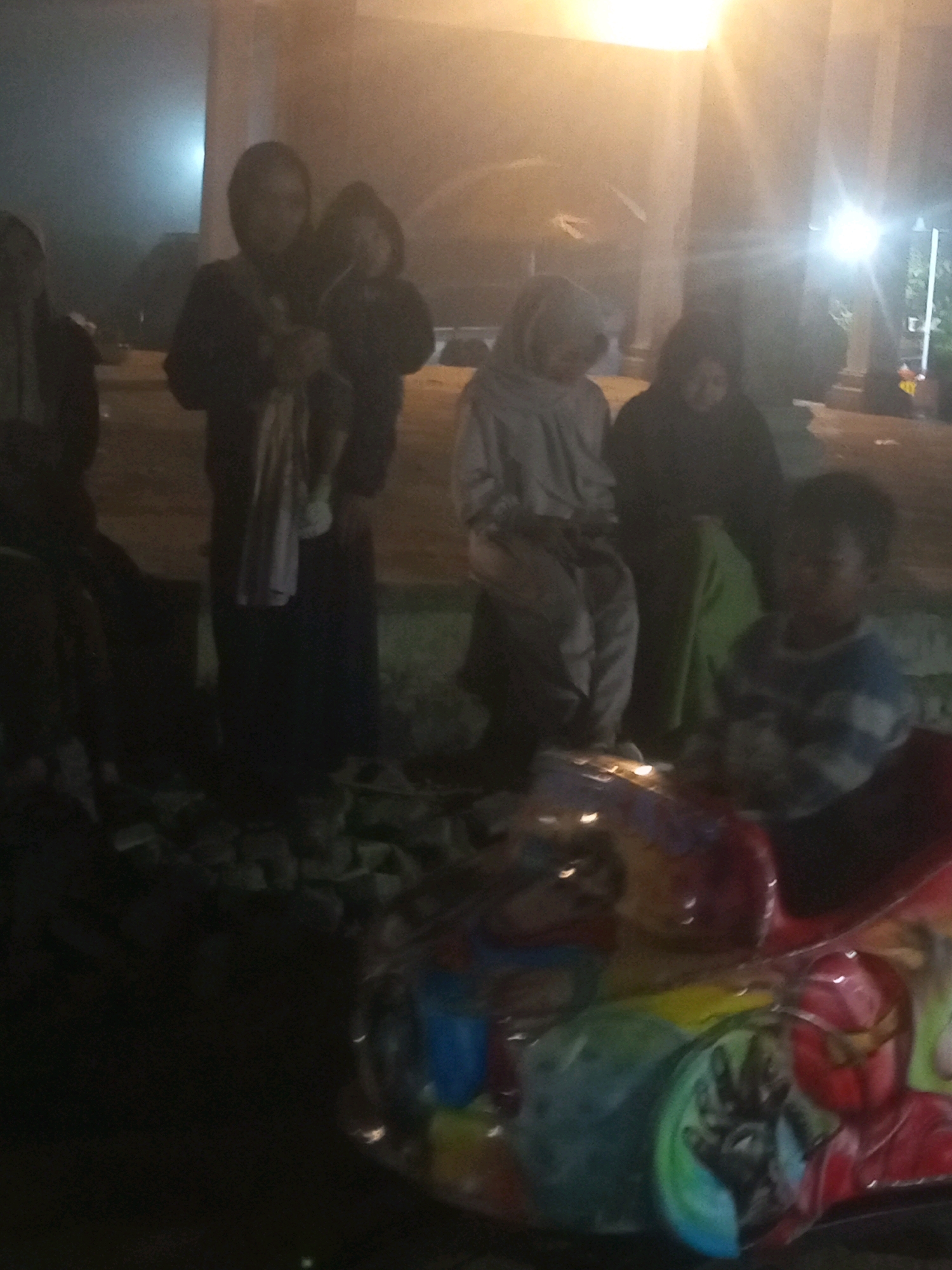 The image depicts a dimly lit outdoor scene in Keseneng, Indonesia, at night. Several individuals are present, appearing to be either standing or sitting. In the foreground, a child is visible, looking towards the viewer. Behind the child, a colorful, possibly decorative, object is present, with figures resembling comic book characters, including what appears to be Iron Man, visible on its surface. The background is obscured by low light and some atmospheric haze, with bright lights suggesting artificial illumination from streetlights or buildings. The ground appears uneven, with scattered debris.