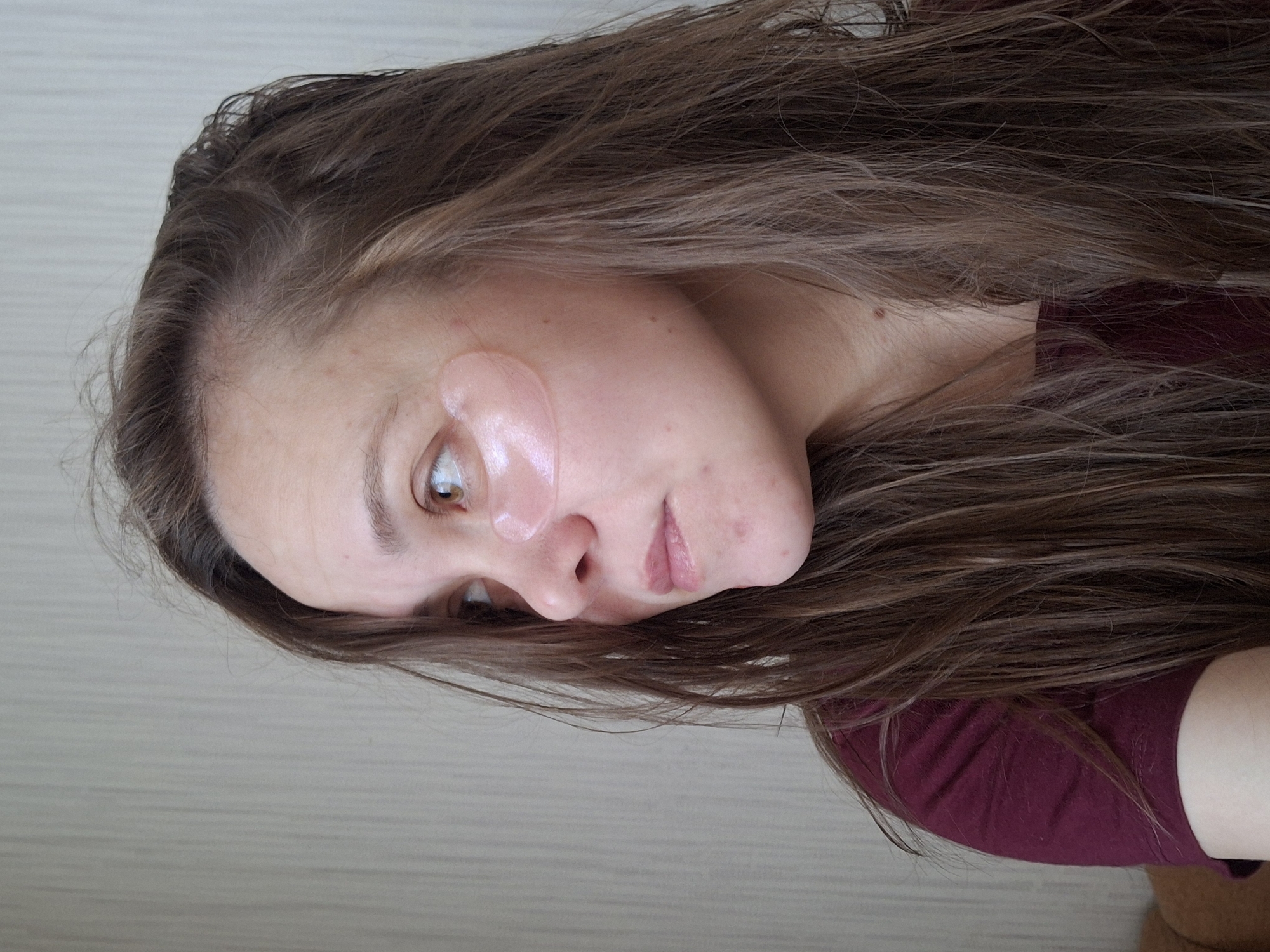 The image shows a close-up, rotated view of a person, likely female, with long, wavy brown hair cascading around her. Her head is tilted, and she is looking away from the camera, downwards and to the right. Two translucent, pearlescent hydrogel eye patches are visible on her left cheekbone, below the eye and extending towards the temple, suggesting a skincare routine or beauty treatment.
Her skin appears fair, with a few small blemishes or freckles visible on her cheek. One brown eye is clearly visible, with a neutral and somewhat reflective gaze. Her lips are partially visible, naturally colored and slightly parted. She is wearing a dark red or burgundy-colored top, partially covered by her hair. Her hair has a natural texture and appears to be slightly damp or recently treated. The hairline along her forehead is visible, showing fine, lighter strands.
The background is a simple, light-colored surface with subtle horizontal lines, indicating an indoor setting, possibly a wall. There are no direct cues about the time of day or weather within the image. The overall impression is one of a quiet, personal moment. No text is visible in the image. The location context provided is Chaikovsky, Russia.