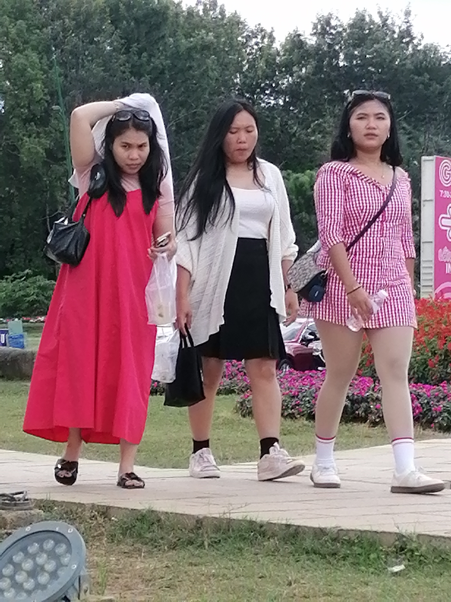Three young Vietnamese women are walking on a paved pathway lined with pink flowers. The woman on the left wears a bright pink dress and holds a black purse and a plastic bag. The middle woman is dressed in a white cardigan over a white top and a black skirt, carrying a black bag and a plastic bag. The woman on the right wears a pink and white checkered dress, white socks, and white shoes, carrying a patterned purse and a water bottle. The background shows lush green trees and a glimpse of a large sign with pink lettering. The scene appears to be in a park or public garden area in Da Lat, Vietnam, given the flora and the general aesthetic.