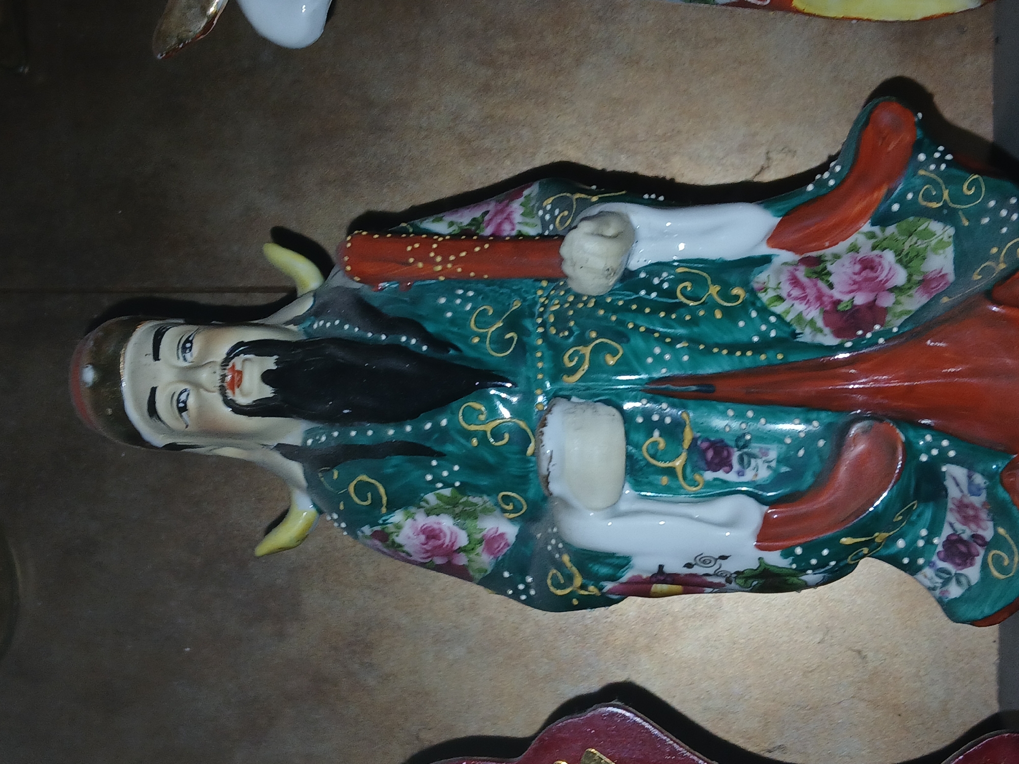 The image features a ceramic or porcelain figurine of an East Asian-style man, lying horizontally on a dark brown surface, possibly wood or tile. The figure is elaborately decorated, wearing a vibrant dark green robe adorned with intricate patterns of pink and white roses, gold swirling motifs, and numerous small white dots. Red accents are visible on the sleeves and collar of the robe.
The man has a distinct long black mustache and goatee, and a calm, serene expression on his face. His head is partially covered by a brown or reddish cap. His light-colored hands appear to be holding a reddish-orange staff or scroll-like object, which is also decorated with small gold dots.
The setting appears to be indoors, likely a display area for decorative items. In the upper background, part of another white ceramic object with a gold rim is visible, and at the bottom foreground, a red and gold decorative piece can be seen. The figurine is stationary, indicating a display rather than an activity or event. There is no visible text in the image. The location context provided is Bengaluru, India, although this is not visually evident from the image itself.