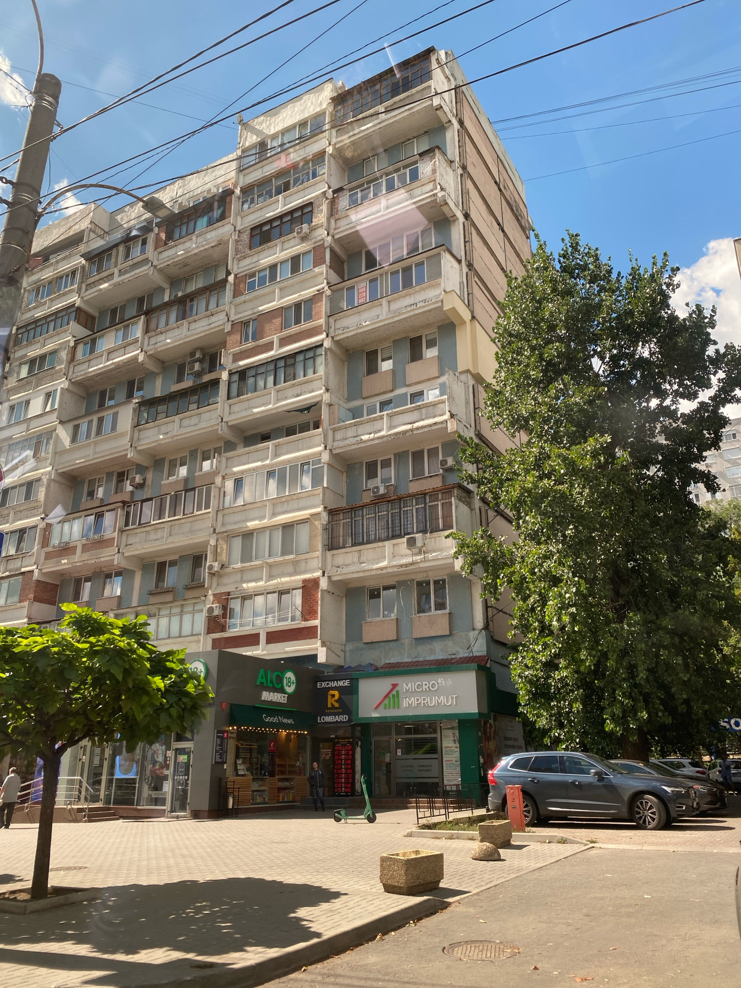 The image shows a wide shot of a street scene in Chișinău, Moldova, on a bright, sunny day. The dominant subject is a multi-story, Brutalist-style apartment building with a light-colored facade, prominent balconies, and rows of windows. The architecture suggests a Soviet-era construction. In the foreground, there is a paved sidewalk and a street. Several trees, one with lush green foliage in the foreground, and another larger one partially obscuring the apartment building on the right, add to the urban greenery.
At the base of the apartment building, there are several storefronts. One is labeled "ALC MARKET" with a "18+" rating and "Good News." Next to it, there is a storefront with "EXCHANGE" and "LOMBARD" signage. A green electric scooter is parked on the sidewalk. Further to the right, a dark gray Volvo SUV and a black car are parked along the street.
In the mid-ground, on the sidewalk, a few people are visible. A man in a light-colored shirt and dark pants walks on the sidewalk in front of the "ALC MARKET." Another person, dressed in dark clothing, is walking near the "EXCHANGE" storefront. A person in a white shirt and dark trousers is seen near the entrance to the "ALC MARKET." The street appears to be made of asphalt, and the sidewalk is paved with light-colored square tiles.
Several electrical wires are visible crisscrossing the sky, and a utility pole is present on the left side of the frame. The sky is blue with scattered white clouds. The overall impression is a typical urban environment in a European city, characterized by residential buildings, commercial establishments, and street infrastructure.