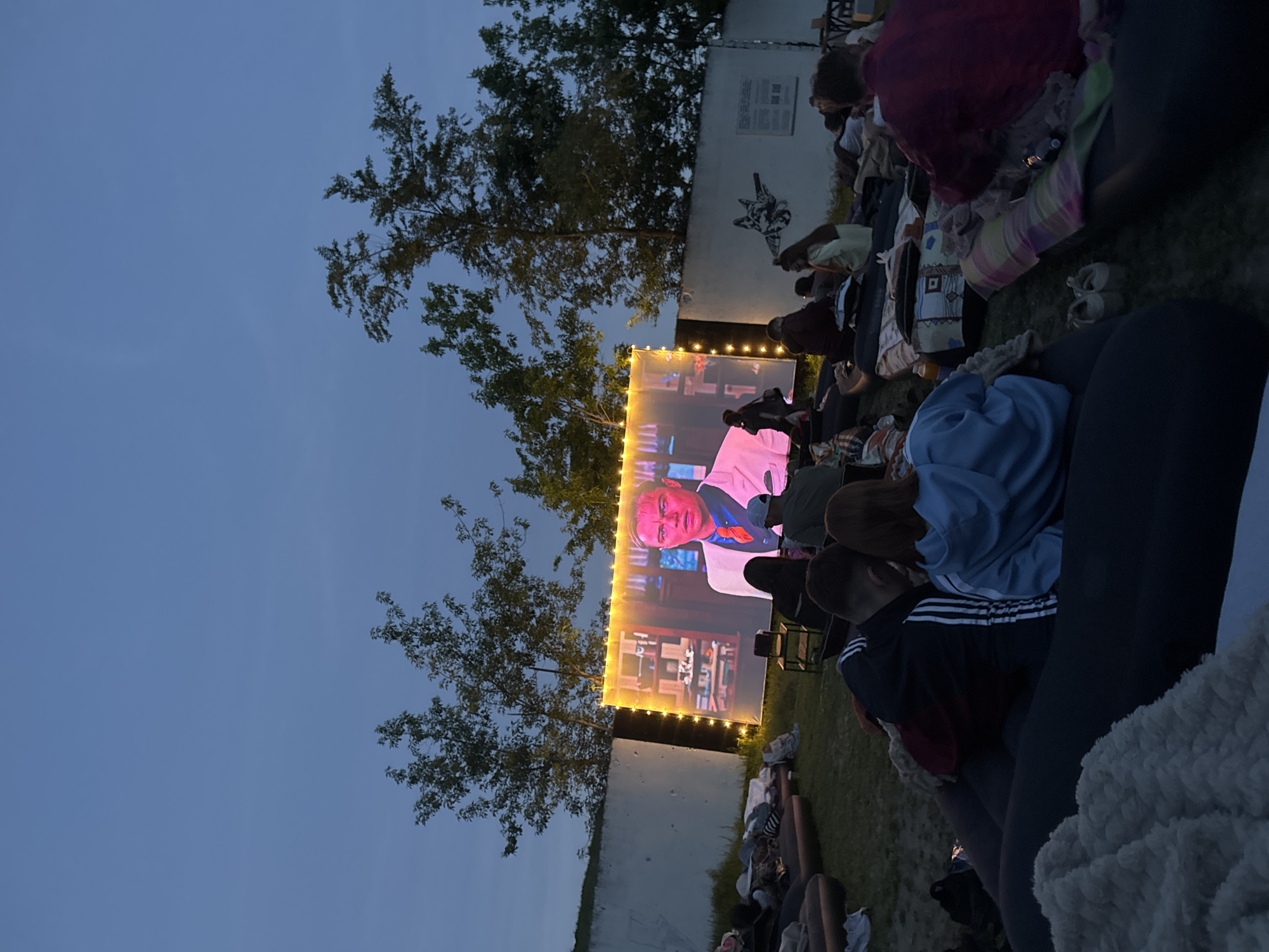 People are gathered outdoors in Saratov, Russia, viewing a movie projected onto a large screen framed with lights. The gathering appears informal, with people sitting on blankets and cushions on the ground. There are trees adjacent to the screen and a wall with artwork. The sky is darkening, indicating it is either dusk or night.
