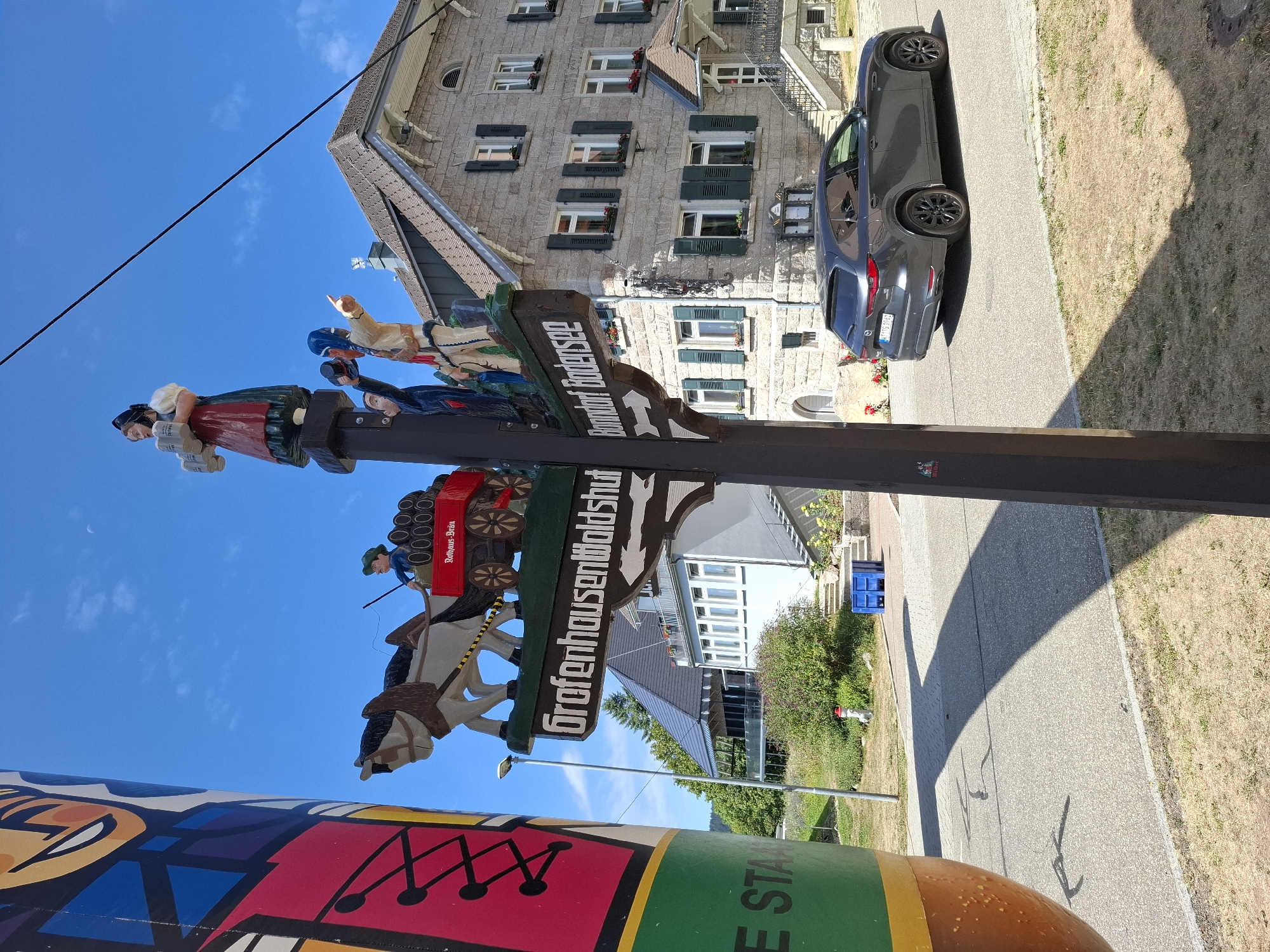 In the foreground of the image, a section of a large, colorful cylindrical object with a pattern of abstract shapes and lines is visible on the left. A wooden signpost stands in the center, featuring carved wooden figures of people and a horse-drawn cart carrying barrels. The sign itself has two arms with text; one reads "Grafenhausen Waldshut" and the other points to "Bonndorf Bodensee". Behind the signpost, a stone building with a tiled roof and multiple windows with green shutters is prominent. A dark gray SUV is parked on the asphalt road in front of the building. The environment appears to be an outdoor setting in a town, with a clear blue sky overhead and some vegetation, including bushes and dry grass, around the buildings and road. The location is identified as Grafenhausen, Germany, by the text on the signpost.