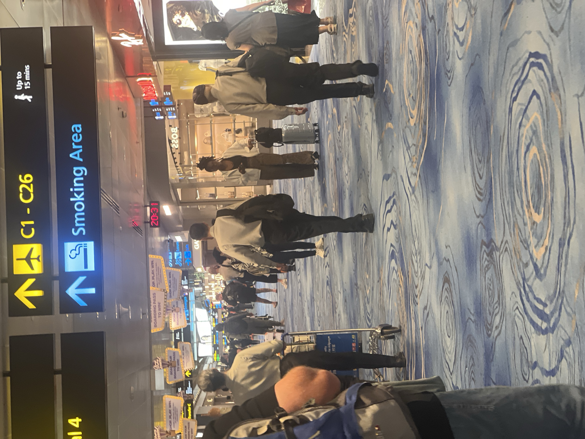 The image depicts a busy airport terminal in Singapore. Overhead signs indicate "C1 - C26" and "Smoking Area." Several individuals are seen walking along a carpeted floor with a swirling blue and gold pattern. Some are pulling luggage or pushing carts. Shops are visible in the background, with displays of merchandise. The lighting suggests it is indoors, likely during the day or evening. The overall scene is that of a typical airport concourse, bustling with travelers.