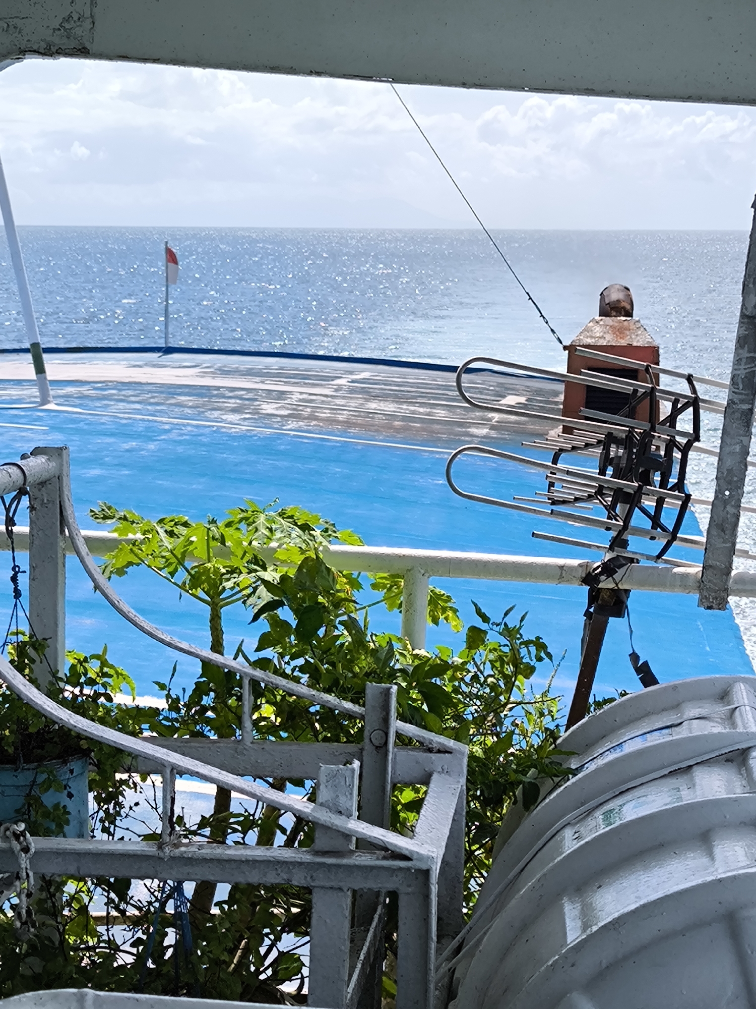 The image presents a view from what appears to be a ship or ferry deck, likely taken in Lembar Selatan, Indonesia, given the reference to the city. The foreground features painted gray metal railings and a structure possibly part of a water tank or life raft storage. Vegetation, including what seems to be a papaya plant, grows in a container near the railing.
Beyond the immediate foreground, the ship's deck is painted a bright blue. A red and white Indonesian flag stands on the deck near the horizon. A small, rusty structure is positioned centrally, with an antenna or similar device extending from it. The background reveals the sea under a partly cloudy sky. The overall scene suggests a working ship with a combination of utilitarian and improvised elements, such as the vegetation.