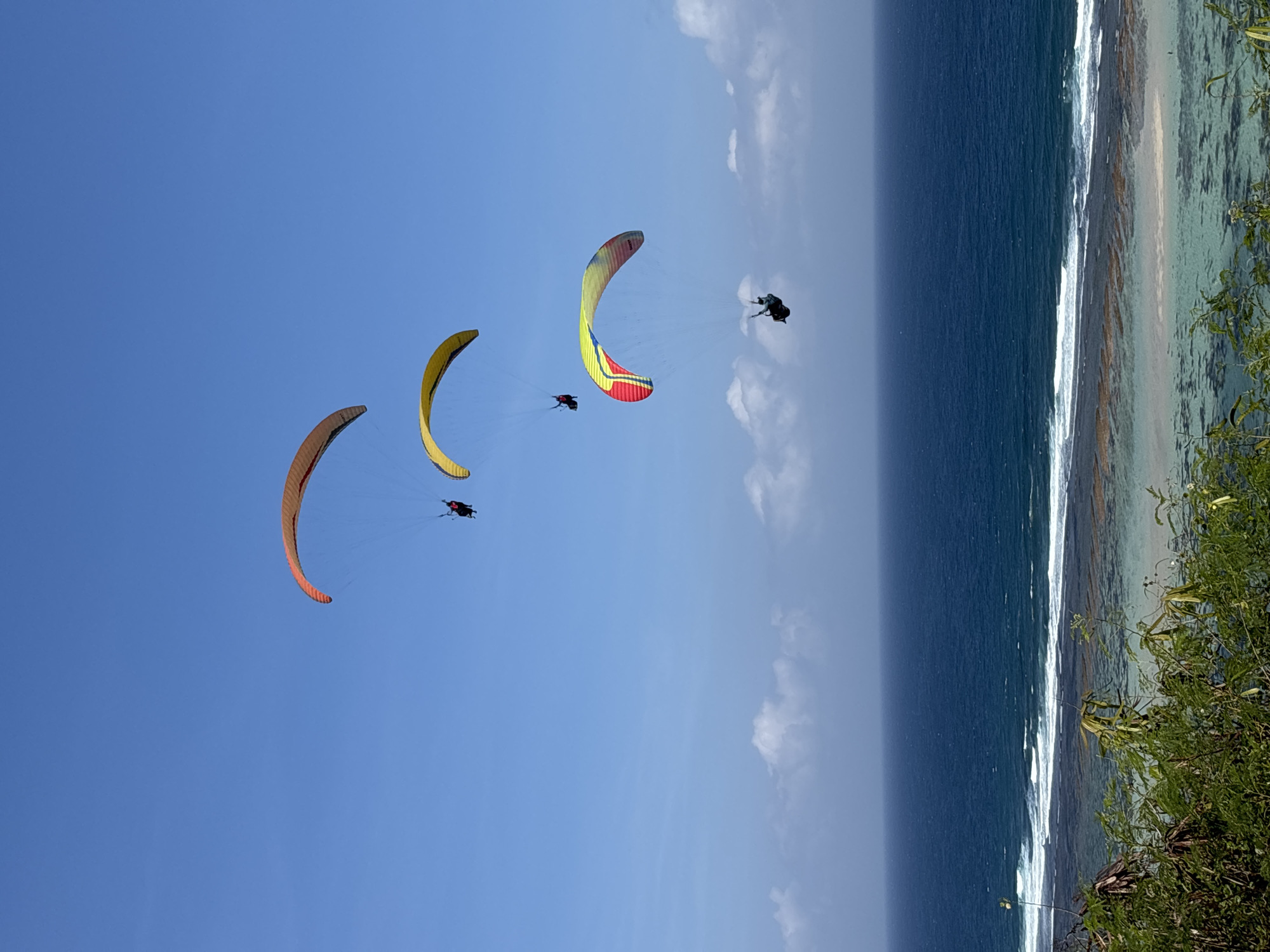 This media file captures three paragliders soaring through a clear blue sky over the ocean, with a coastline visible in the background. The paragliders, each with a colorful parachute, appear to be enjoying a sunny day. The paraglides have distinct colors: one is yellow, another is a red and yellow combination, and the third is orange. One paraglider is clearly visible with a dark parachute and a rider wearing a red and black suit.
The scene is set by the vast expanse of the ocean, transitioning to a shallow, turquoise water area near the shore where waves are breaking. The coastline shows a sandy beach and rocky formations. The time of day appears to be midday, given the bright sunlight casting clear shadows and illuminating the scene. The weather seems to be clear and pleasant, ideal for paragliding.
The paragliders are engaged in the activity of paragliding, a recreational activity where individuals glide through the air suspended by a fabric wing. The image conveys a sense of freedom and exhilaration associated with the sport. The location cues, such as the tropical coastline and the type of waves, suggest a beach destination. There is no visible text in the media file. The overall mood is one of adventure and enjoyment of the natural beauty of the location.