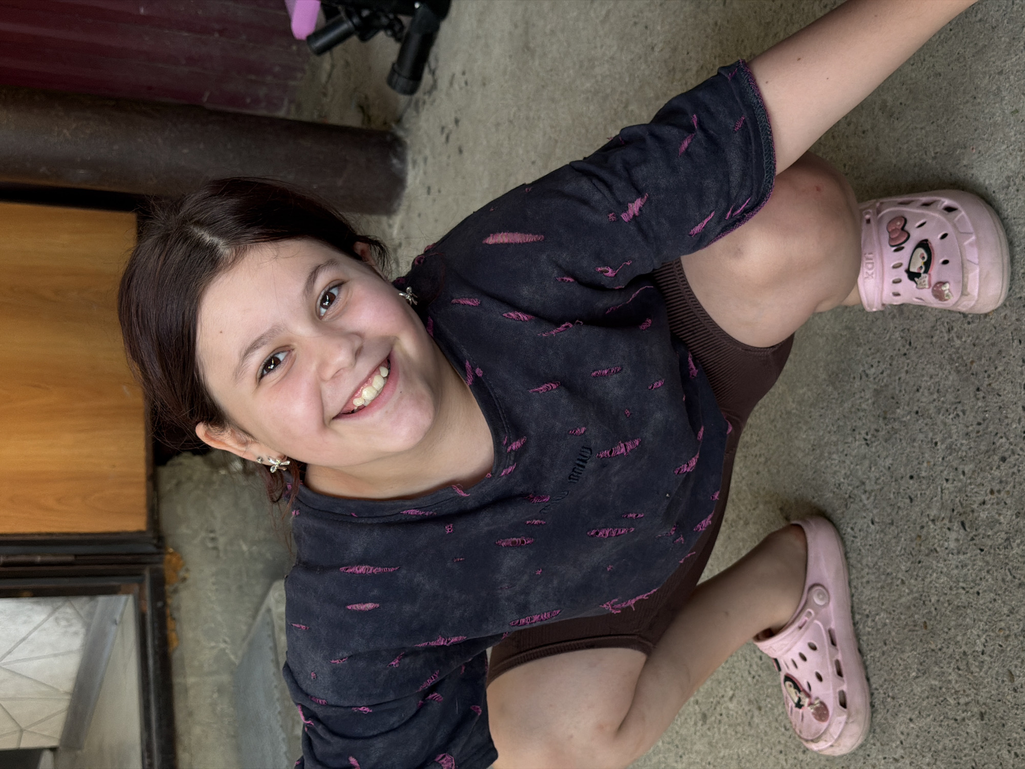 This media file shows a young girl crouching outdoors. She has dark brown hair and is smiling brightly at the camera. She is wearing a dark blue t-shirt with pink distressed detailing and brown shorts. On her feet, she has pink slides with cartoon characters. The setting appears to be a concrete area, possibly a patio or backyard, with some wooden elements and what looks like a metal structure in the background. The lighting suggests it is daytime. The girl's expression conveys happiness and playfulness. No visible text is present on the girl's clothing or surroundings.