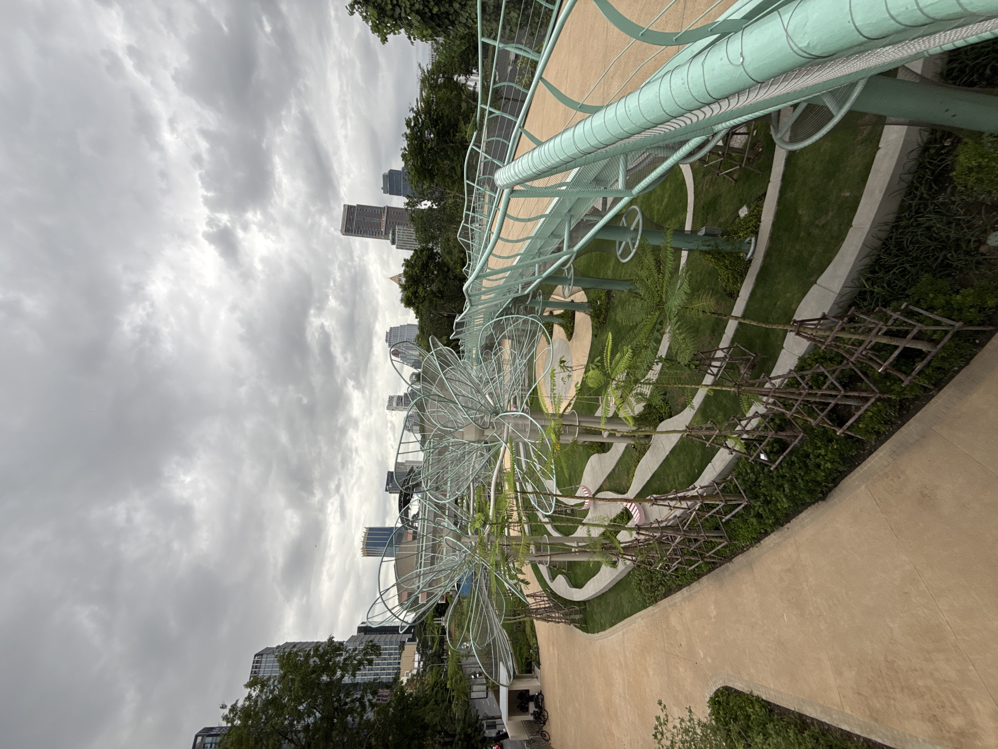 This is a medium-angle, eye-level, full shot of a park area in Bangkok, Thailand, on a cloudy day. The foreground features a beige-colored pedestrian walkway curving to the left. To the right, a light green, elevated walkway with a curved railing made of metal mesh and pipes is visible. The walkway is supported by a series of light green, tubular structures.
In the middle ground, the park displays a landscaped area with manicured green grass, winding pathways, and several young trees with wooden supports. Large, intricate wire sculptures resembling stylized flowers or trees are a prominent feature. Two pink swings hang from one of these sculptures.
In the background, several modern, tall buildings are visible against a cloudy sky. A tree with dark green foliage is on the far left. On the far right, more dense green trees are present. On the far left, at ground level near the pedestrian walkway, there are two motorcycles parked outside a small, open-sided structure. No people are clearly visible, but their presence is implied by the motorcycles and the park's design. The overall impression is a modern urban park designed with artistic elements and greenery, set against a backdrop of city architecture.