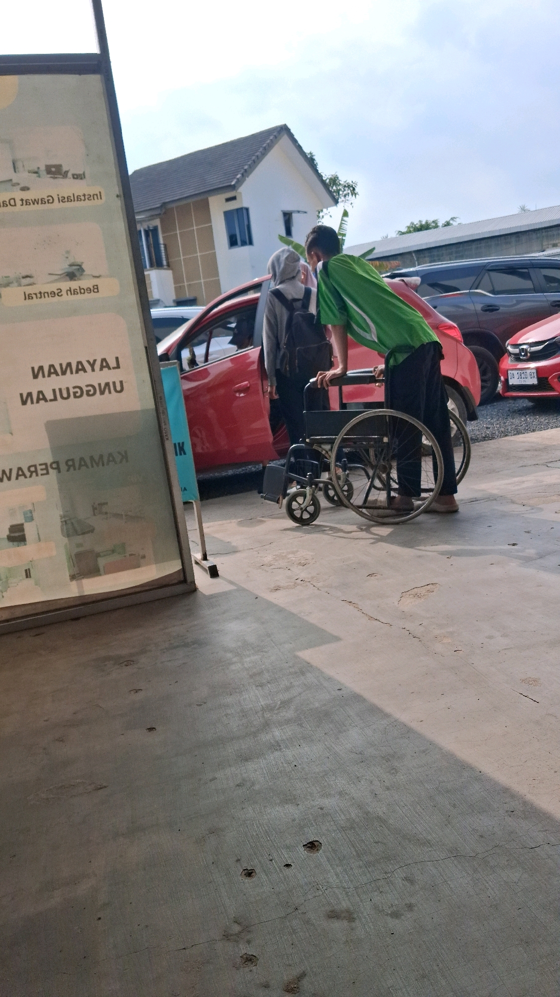 In Banjarbaru, Indonesia, two individuals, one wearing a green shirt and the other a grey hoodie and headscarf, are near a red car. The person in the green shirt appears to be pushing a wheelchair. The scene suggests a public area, possibly a parking lot or an entrance, with a building and other vehicles visible in the background. The lighting indicates it is daytime. A sign with text in an Indonesian language is positioned to the left of the frame.